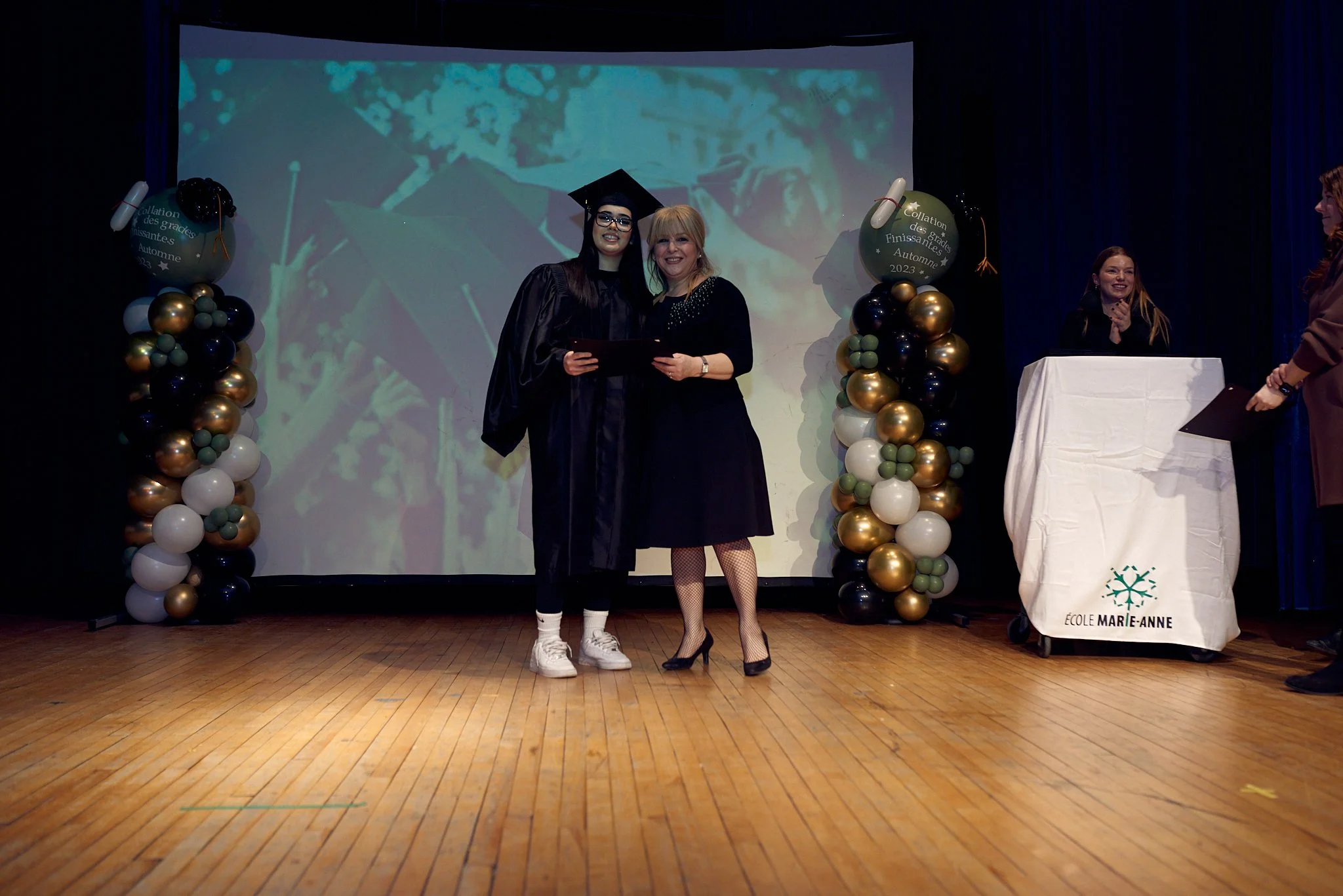 Une cérémonie de remise de diplôme à l'école Marie-Anne, avec une étudiante en costume de graduation recevant son diplôme d'une femme vêtue de noir, sur une scène décorée avec des ballons.