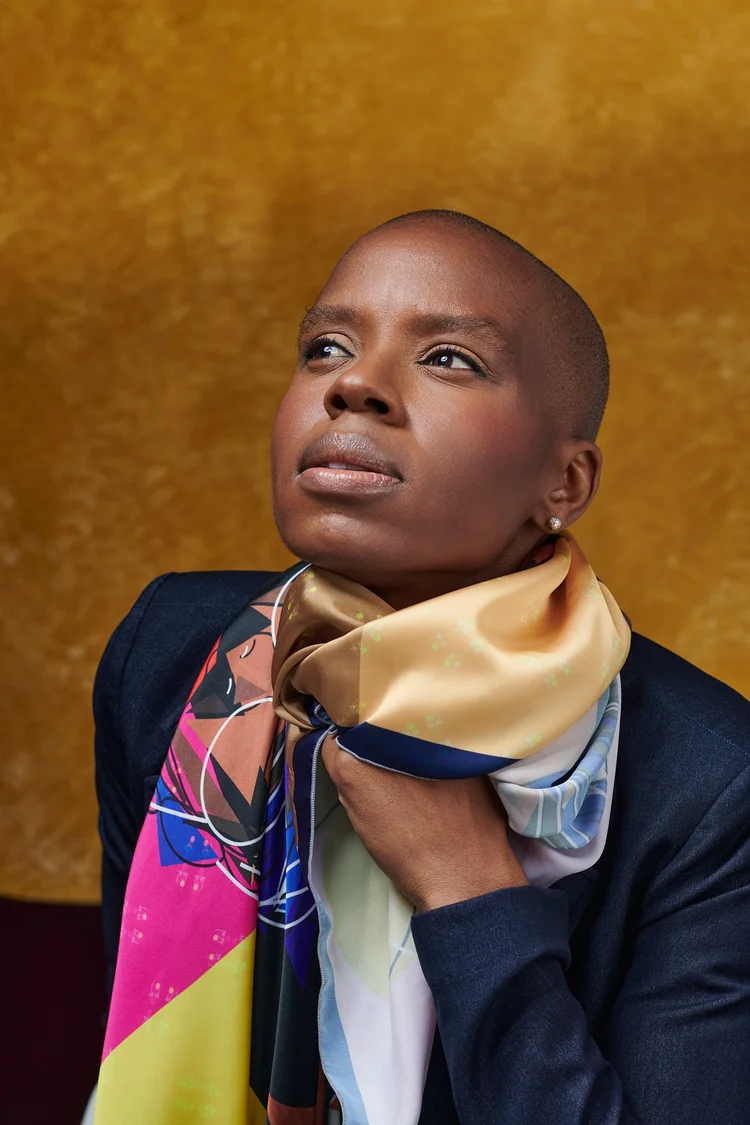 Portrait d'une femme noire portant une veste sombre, un foulard en soie beige avec des motifs, et une boucle d'oreille en diamant. Son regard est tourné vers la gauche, avec une expression sérieuse. L'arrière-plan est doré.