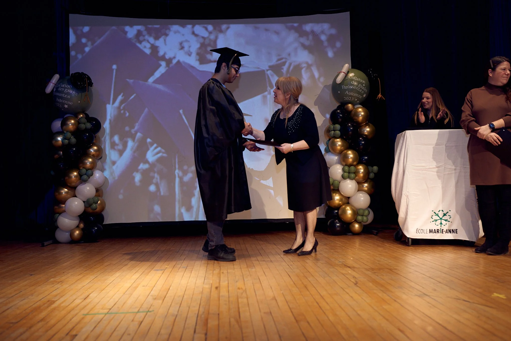 Une cérémonie de remise de diplôme dans une école with balloons colorés en noir, blanc, doré et vert, et une grande projection en arrière-plan montrant des étudiants diplômés portant leur chapeau de graduation.