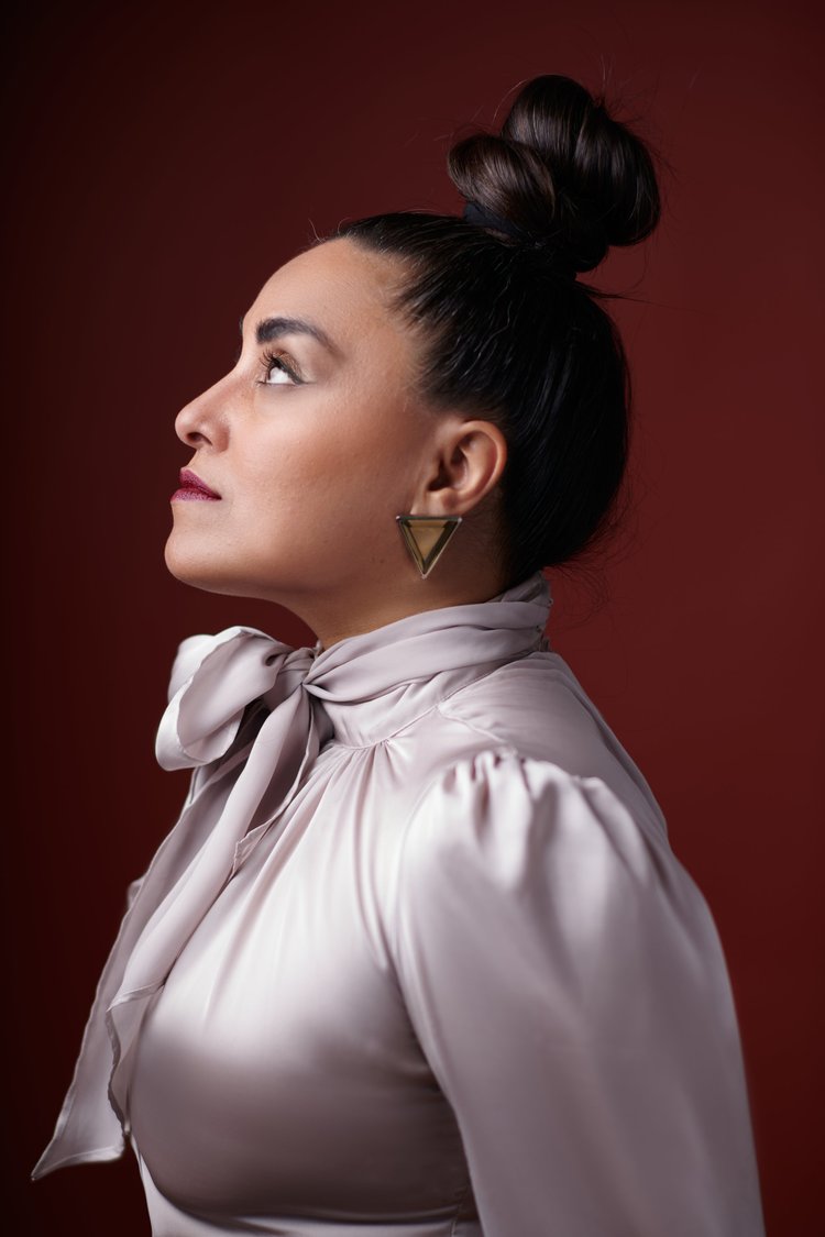 Portrait d'une femme coiffée d'un chignon bun, portant un haut en satin beige avec un nœud au col, une boucle d'oreille triangulaire dorée, regardant vers la gauche, avec un fond rouge brûlé.