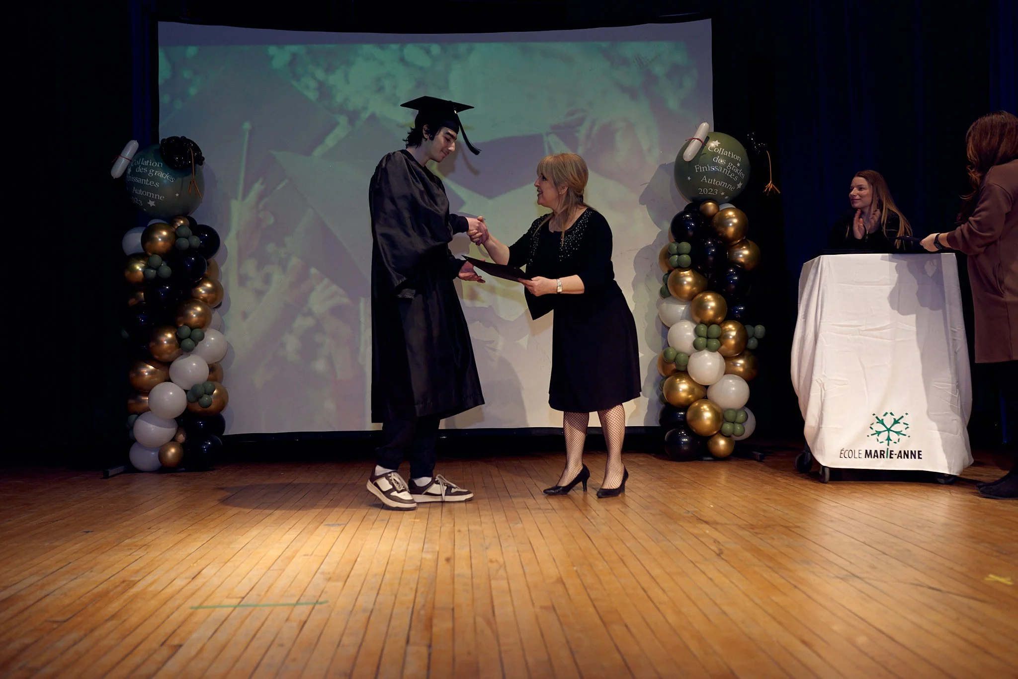 Un étudiant en costume de graduation reçoit son diplôme lors d'une cérémonie de fin d'études à l'école Marie-Anne, avec des ballons décoratifs autour