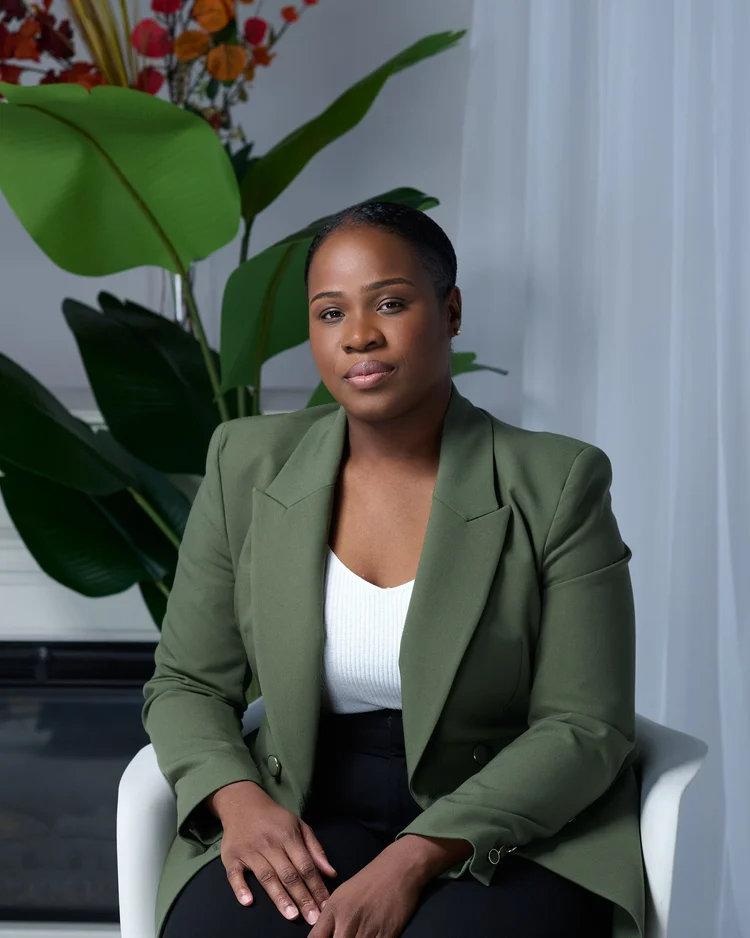 Femme assise portant un blazer vert olive sur un haut blanc, mains croisées sur les genoux, devant un rideau clair et une plante verte.