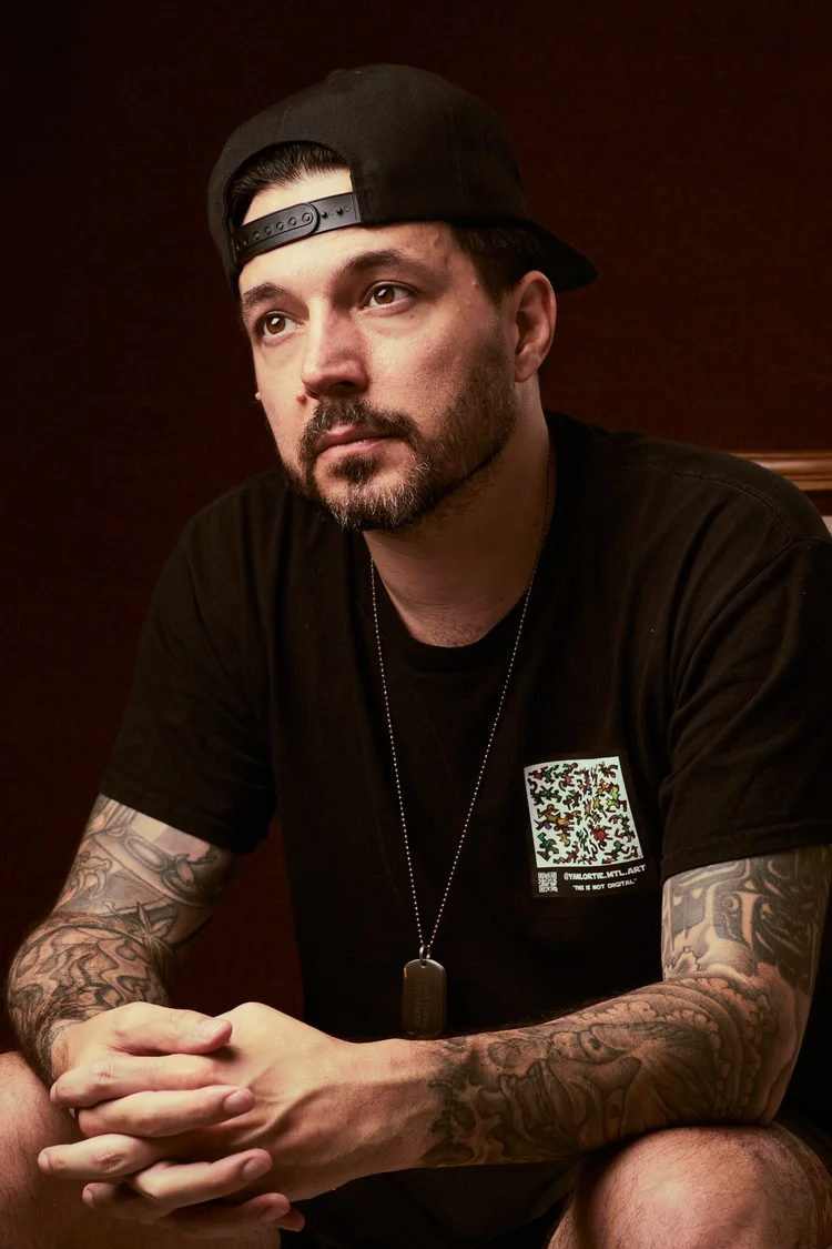 Homme aux avant-bras tatoués, casquette noire portée à l'envers, t-shirt noir et collier pendentif, assis, mains jointes, regard tourné vers le côté.
