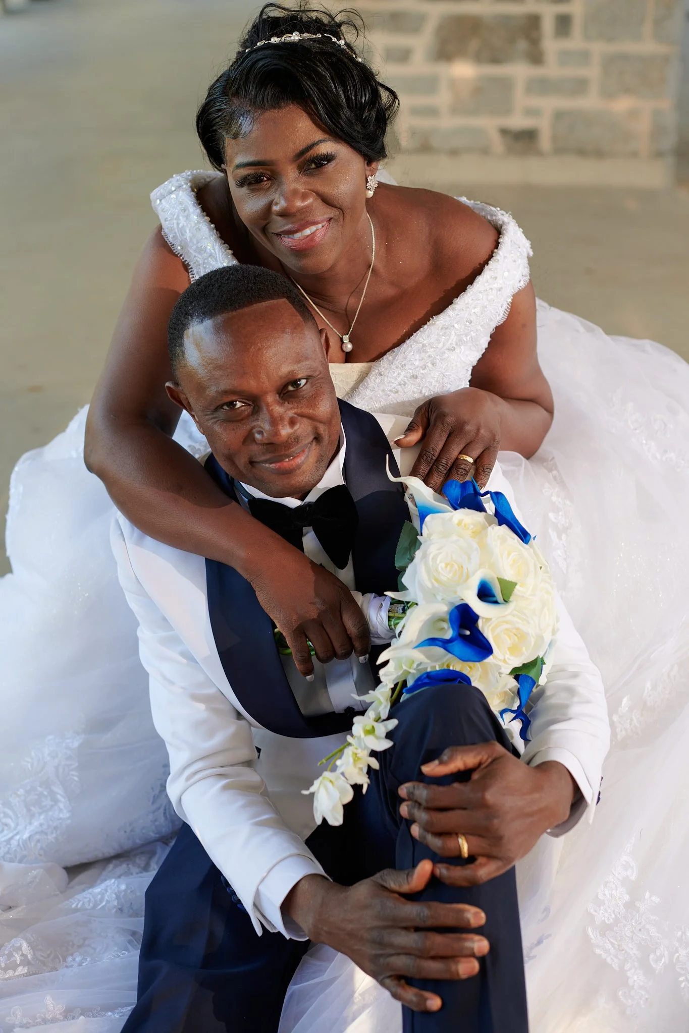 Un couple de mariés pose pour une photo de mariage, la femme en robe blanche et le marié en smoking, avec un bouquet de fleurs blanches avec des touches de bleu.