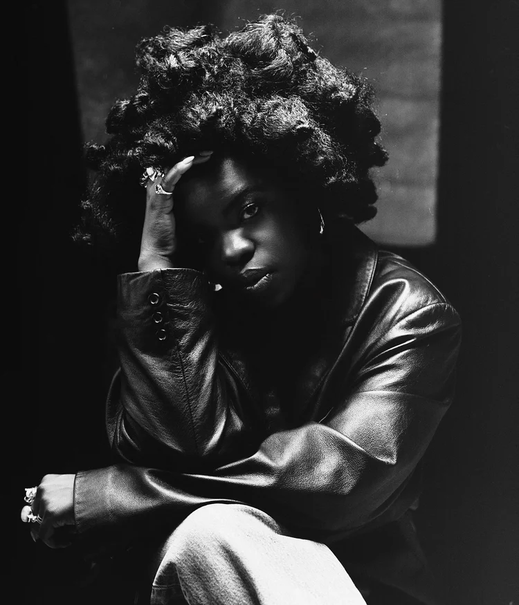 Une femme noire avec des cheveux bouclés et frisés, portant une veste en cuir, pose avec sa main sur son visage, dans une photographie en noir et blanc.