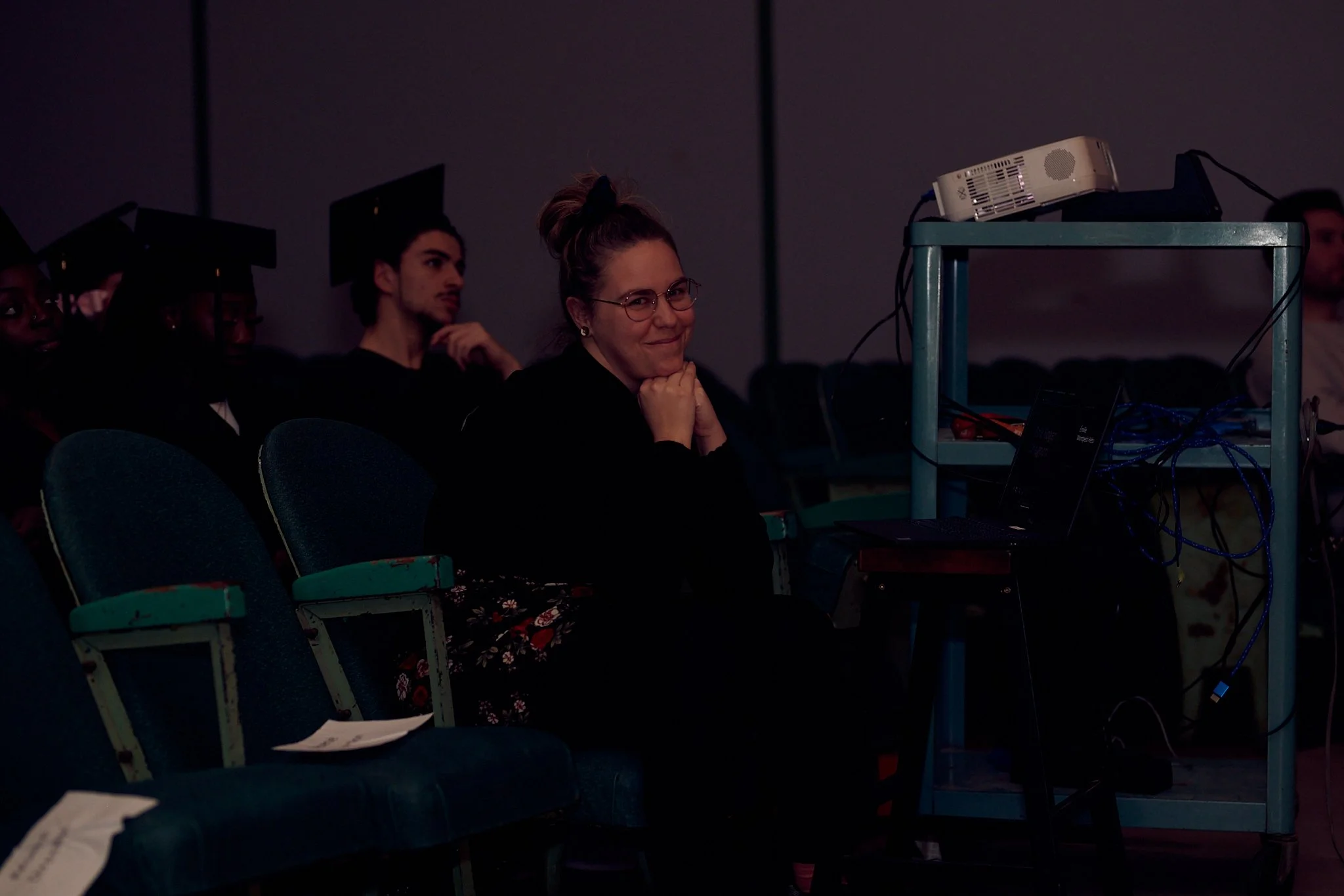 Une femme souriante et assise dans une salle de conférence, portant des lunettes et regardant vers la caméra. Elle est entourée de personnes avec des chapeaux de graduation, assises dans des fauteuils bleus.