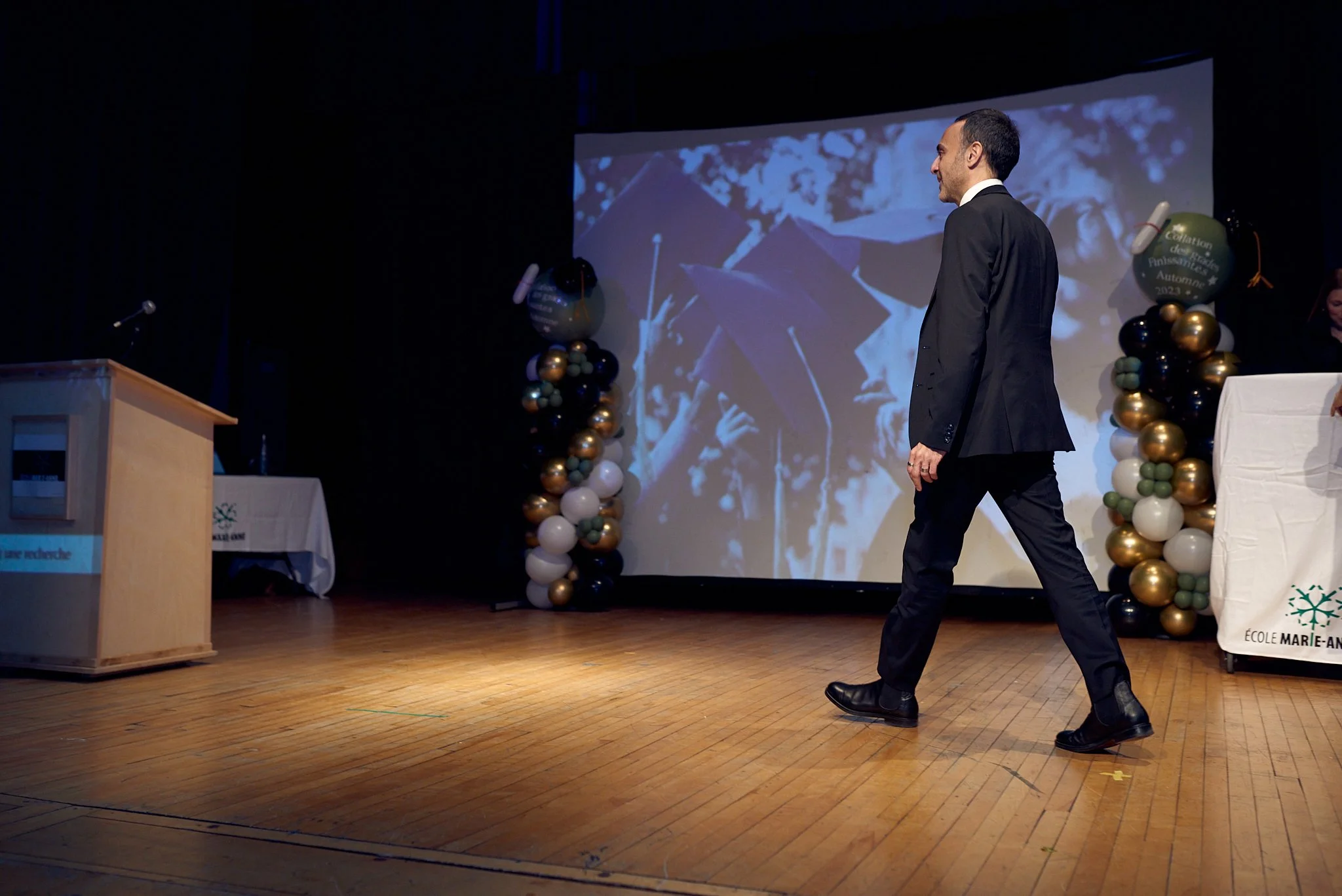 Un homme en costume noir marche sur scène lors d'une cérémonie, avec un écran en arrière-plan montrant des diplômés portant leur toge et chapeau de graduation. La scène est décorée de ballons colorés et il y a un pupitre et une table à droite.