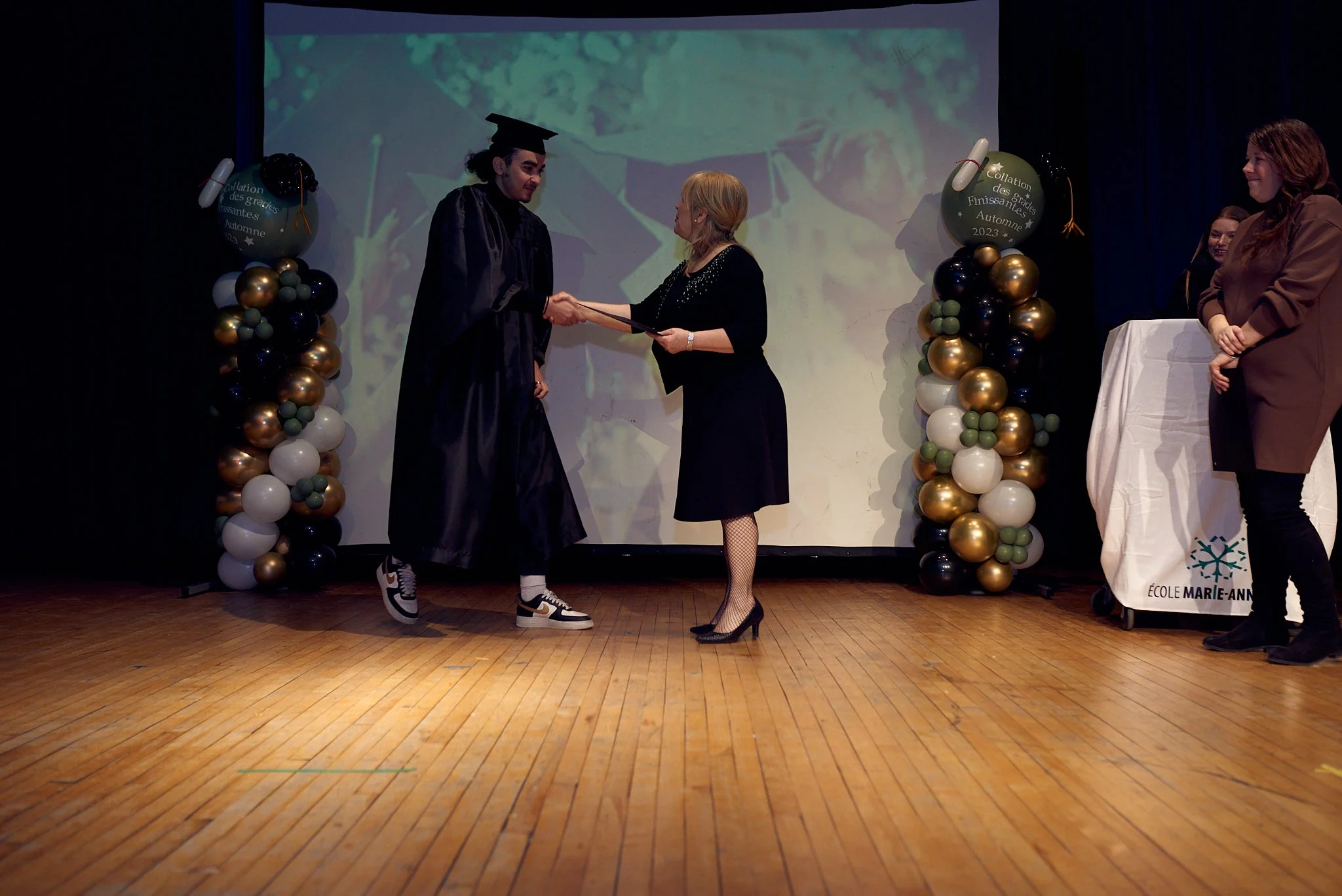 Un diplôme est remis à un étudiant lors d'une cérémonie de remise des diplômes à l'école Marie-Anne, avec des décorations de ballons dorés, noirs, blancs et verts.