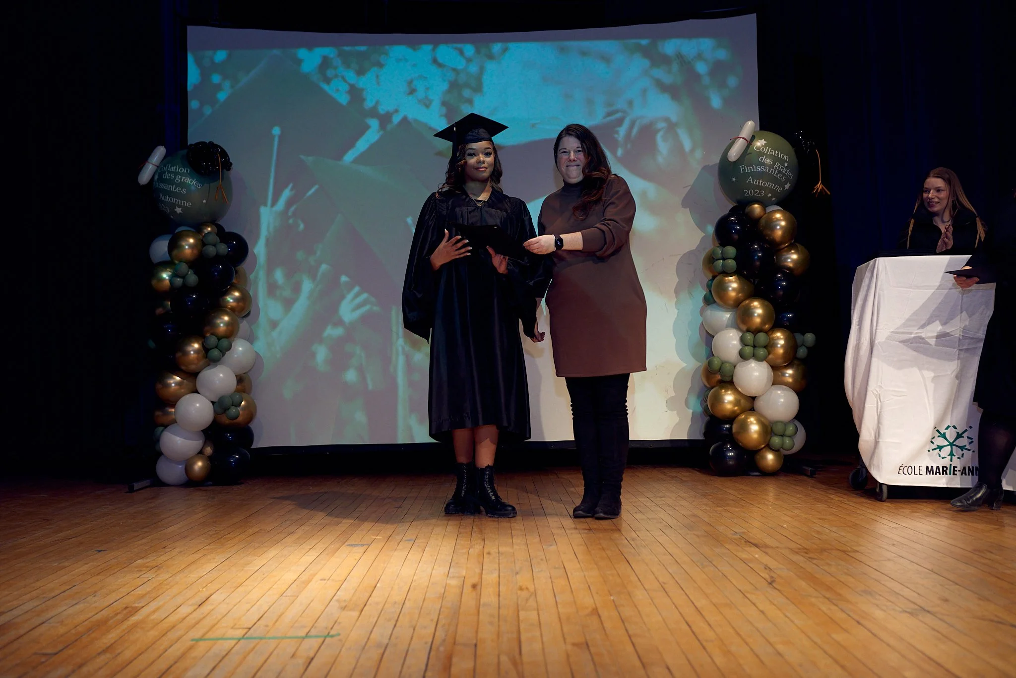 Une cérémonie de remise de diplôme avec une étudiante en costume de graduation recevant un diplôme d'une femme sur scène, entourée de décorations de ballons, lors d'une graduation d'automne.