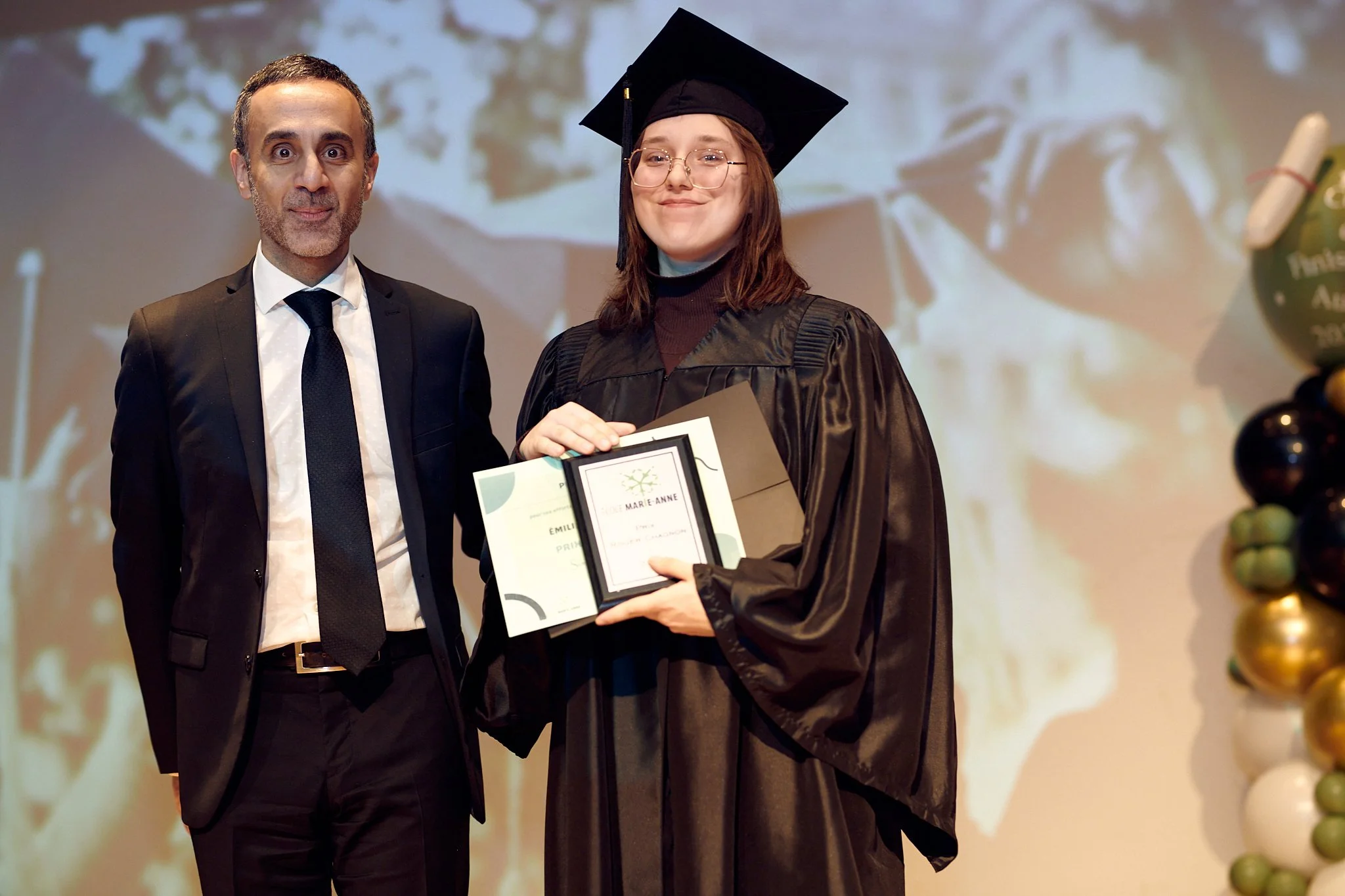 Une personne en robe de graduation tenant un certificat, posant avec un homme en costume lors d'une cérémonie de remise de diplômes.
