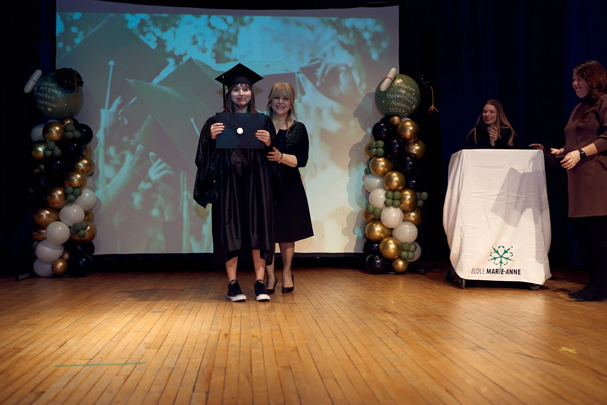 Une cérémonie de remise de diplôme à l'école Marie-Anne. Une jeune diplômée en robe de graduation et chapeau, souriante, posant avec une femme en costume avec un ruban blanc. En arrière-plan, un écran avec une image de graduation. À droite, deux femm