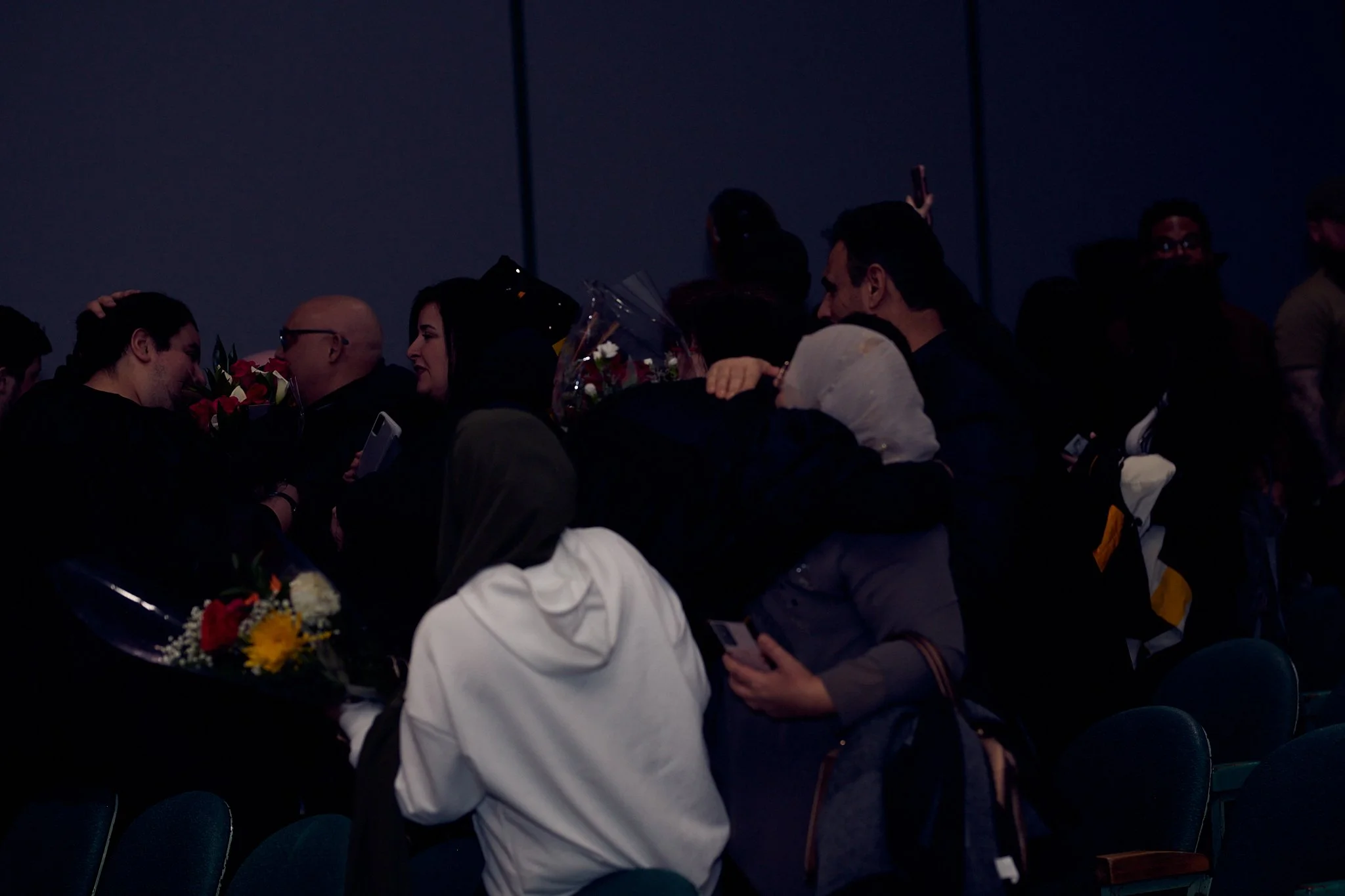 Groupe de personnes échangent des fleurs et des saluts lors d'un événement en intérieur, sous un éclairage sombre.