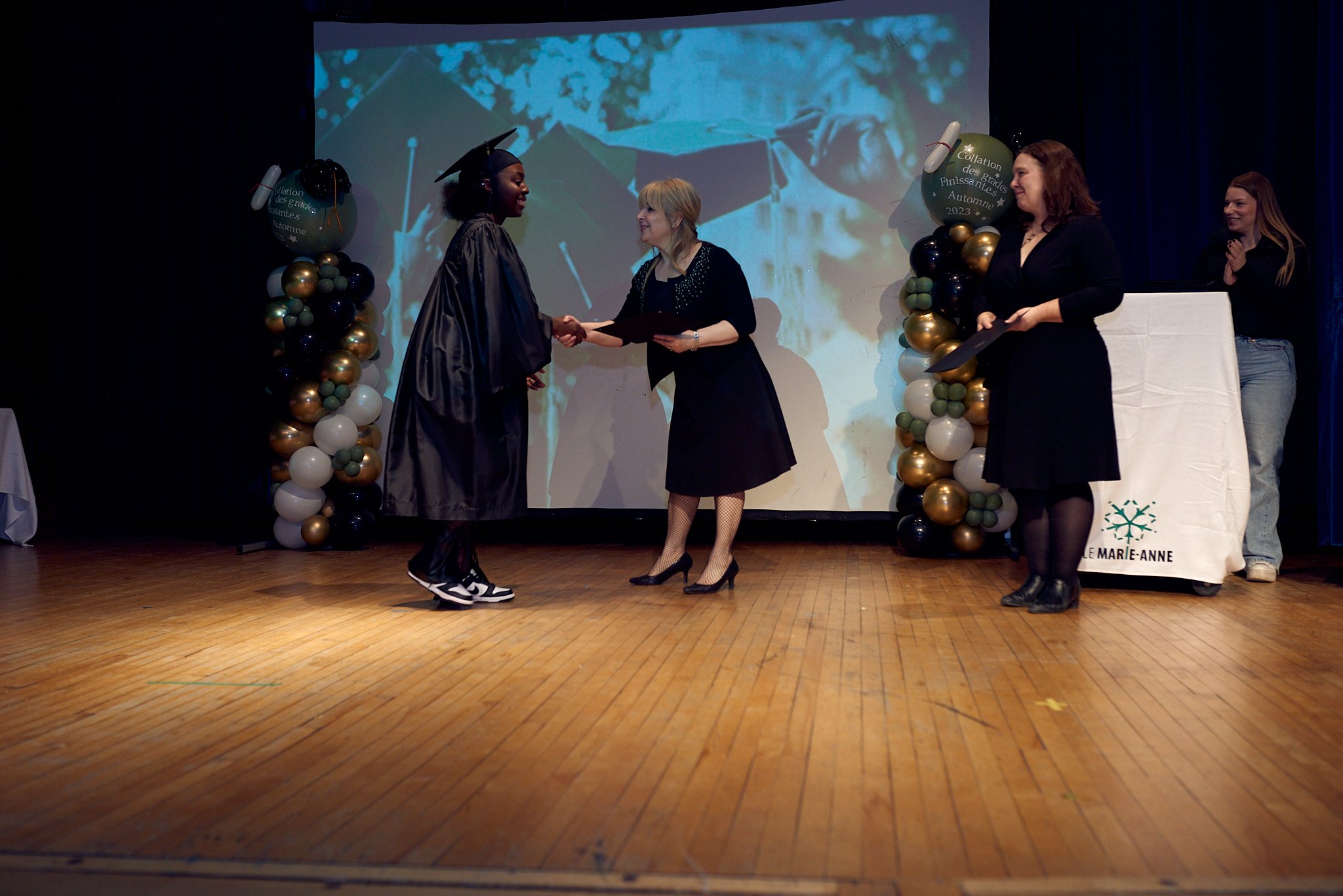 Une cérémonie de remise de diplôme avec une personne diplômée en graduation et un personnel qui lui remet un certificat sur scène, décorée de ballons et d'une bannière.