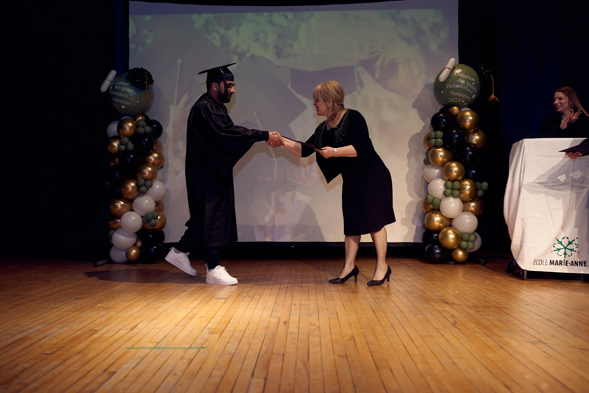 Un diplômé en robe de graduation serre la main d'une femme en costume lors d'une cérémonie de remise de diplôme, avec des décorations de ballons dorés, blancs, gris et noirs en arrière-plan.