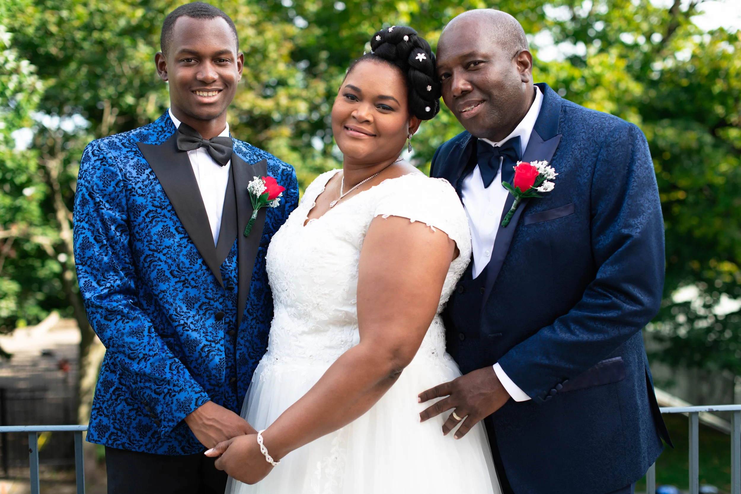 Groupe de quatre personnes lors d'une cérémonie de mariage, deux hommes en costumes bleus avec boutonnières rouges, une femme en robe de mariée blanche, tous souriants dans un parc avec des arbres en arrière-plan.