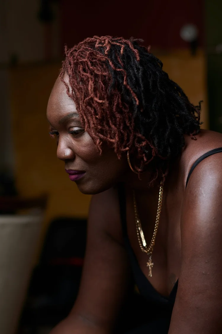 Une femme avec des tresses rouge foncé et noires regarde vers le bas, portant des boucles d'oreilles en or, un collier orné d'une croix, et une robe noire à bretelles, dans un environnement intérieur peu éclairé.