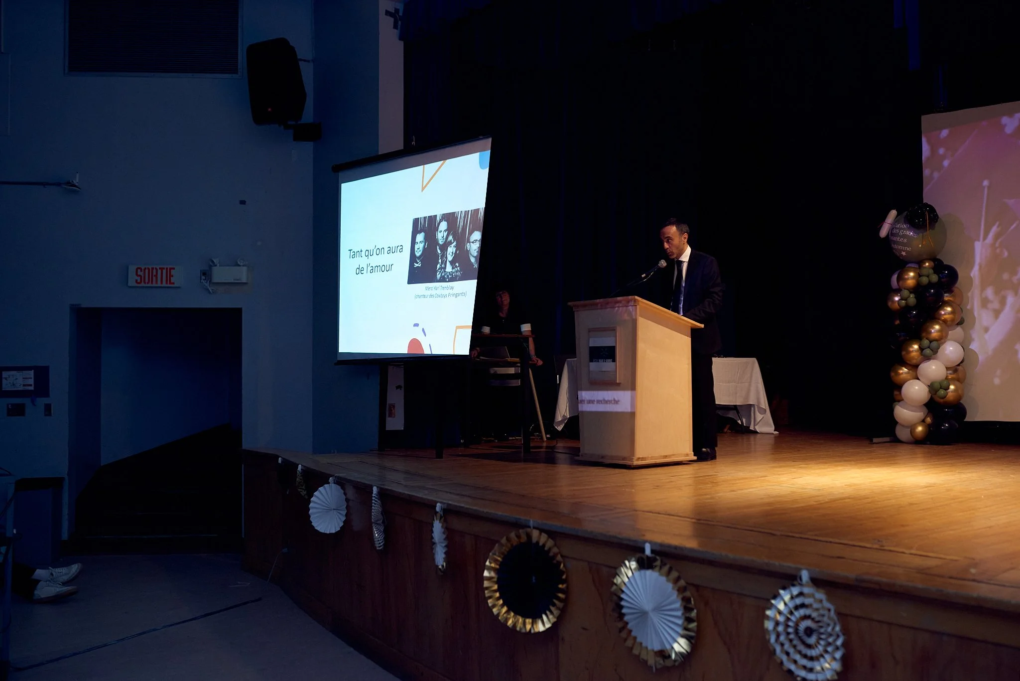 Un homme en costume noir parlant à un micro derrière un pupitre lors d'une présentation sur scène. À gauche, un écran affiche une diapositive avec des images de personnes et du texte français. La scène est décorée avec des ballons et des guirlandes, 