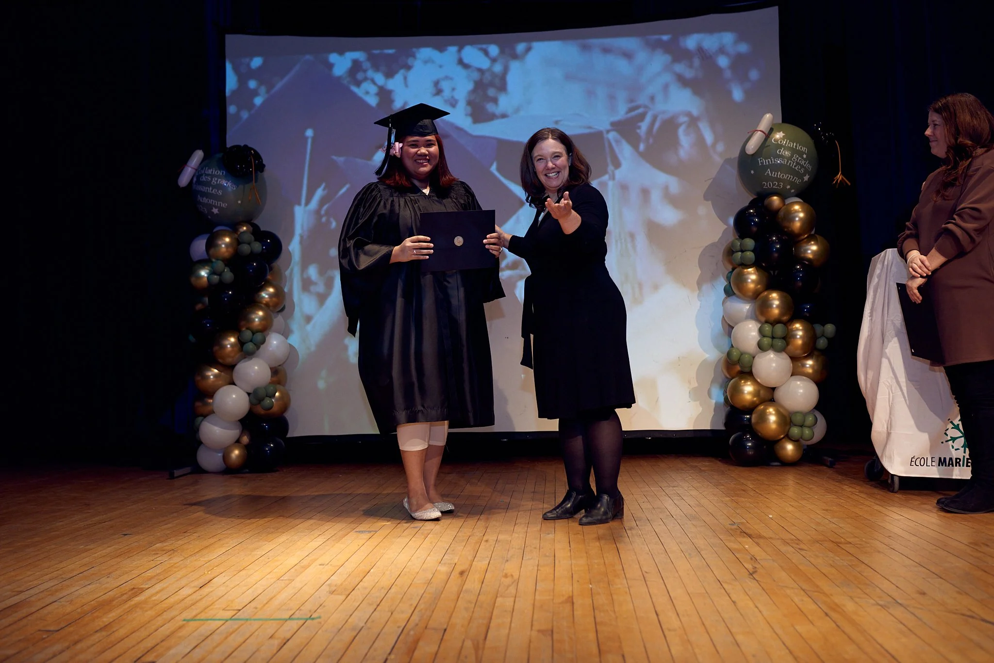 Une cérémonie de remise de diplôme sur scène avec deux femmes, une en robe de graduation et une autre en vêtement noir, souriantes. Décorations avec ballons et une projection en arrière-plan.
