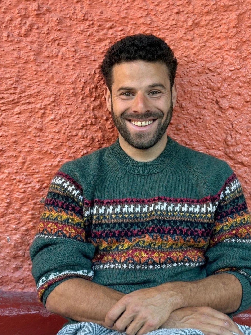 A man with short curly dark hair and a beard smiling, sitting against a textured red-orange wall, wearing a Christmas sweater with colorful patterns.