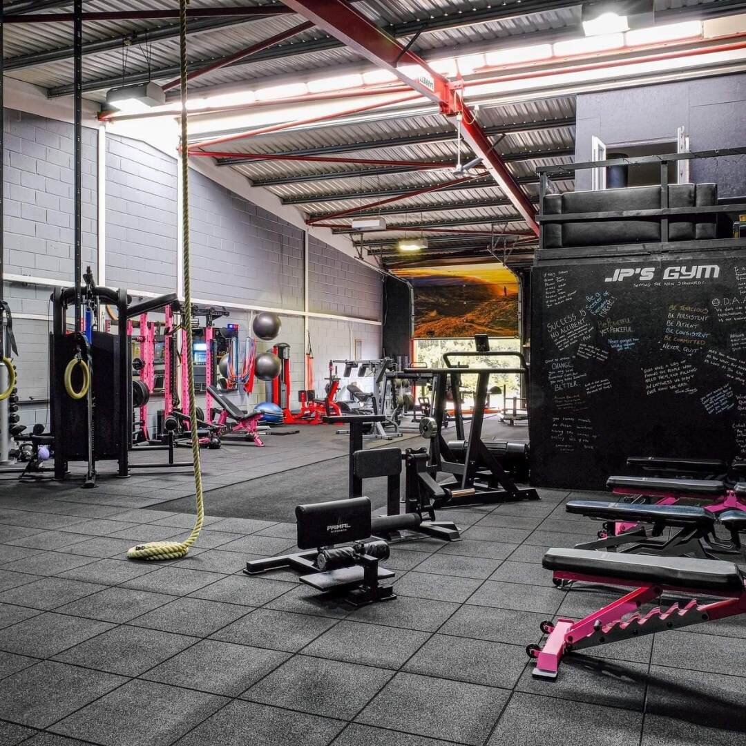 Interior of a gym with various fitness equipment, including weight benches, exercise machines, ropes, and exercise balls. The walls display motivational quotes. The space has a concrete floor with rubber mats, and a high metal ceiling with bright lig