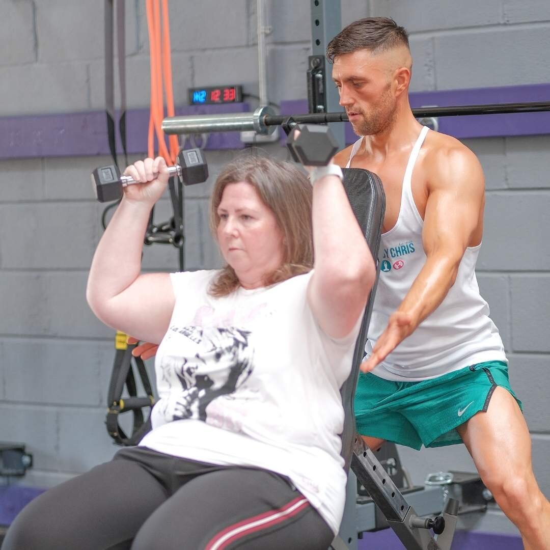 A woman lifting dumbbells seated in a gym, assisted by a male trainer wearing a tank top and shorts.