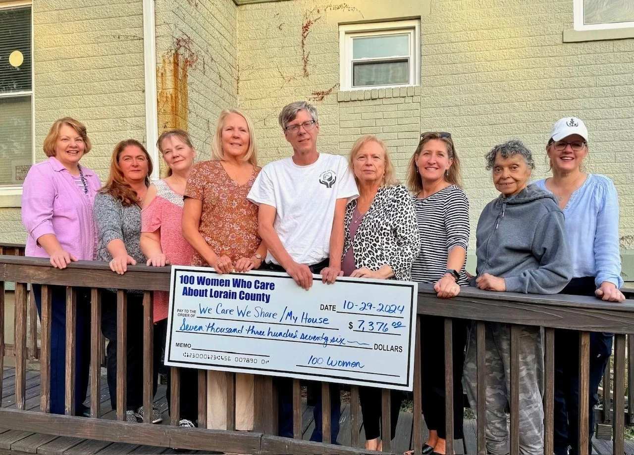 A group of ten women and one man standing on a wooden deck holding a large check for $7,376 made out to 'We Care We Share / My House' from 100 Women Who Care About Lorain County, dated October 29, 2024.