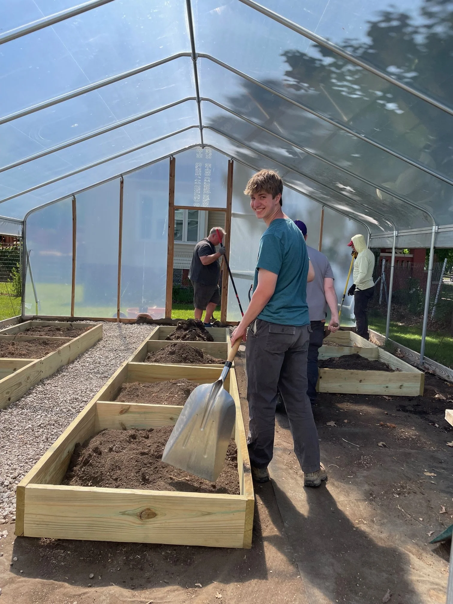 People working in a greenhouse, planting soil in wooden garden beds, with one young man smiling at the camera.