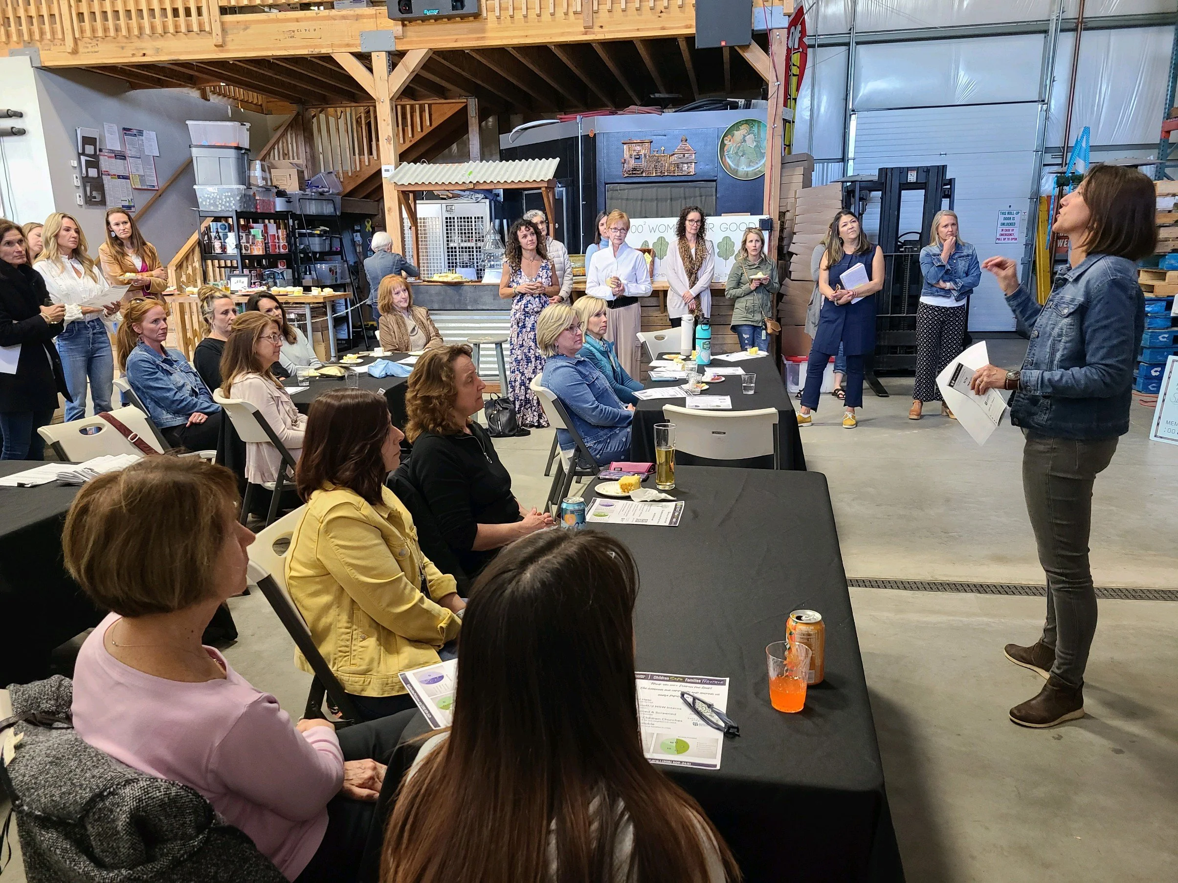 A woman in a denim jacket is giving a presentation to a group of women in an industrial-style room with wooden accents and storage shelves. The women are seated and standing, listening attentively; some have drinks and papers in front of them.