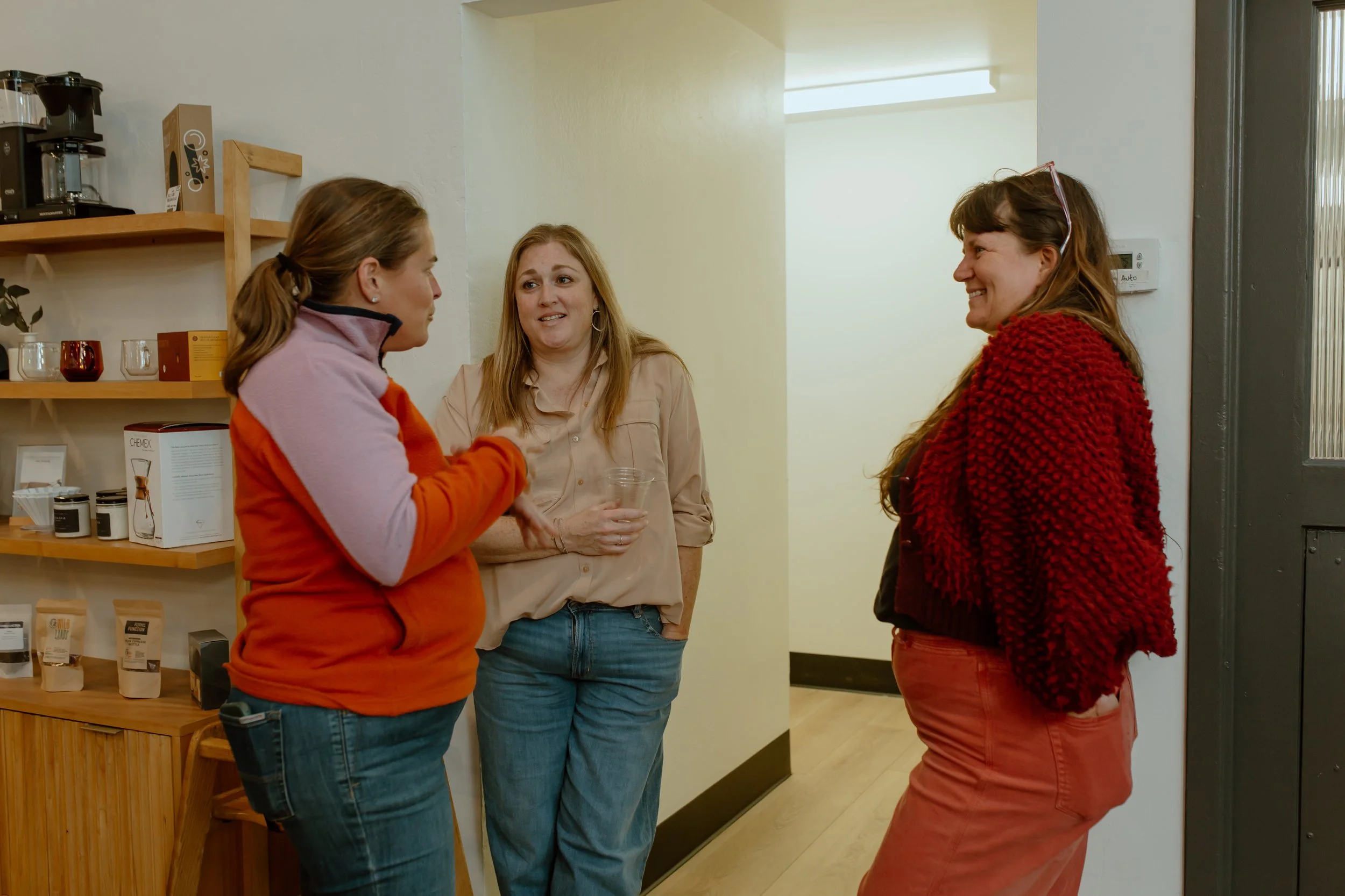 Three women converse in a cozy coffee shop, standing near wooden shelves with coffee equipment and cups.