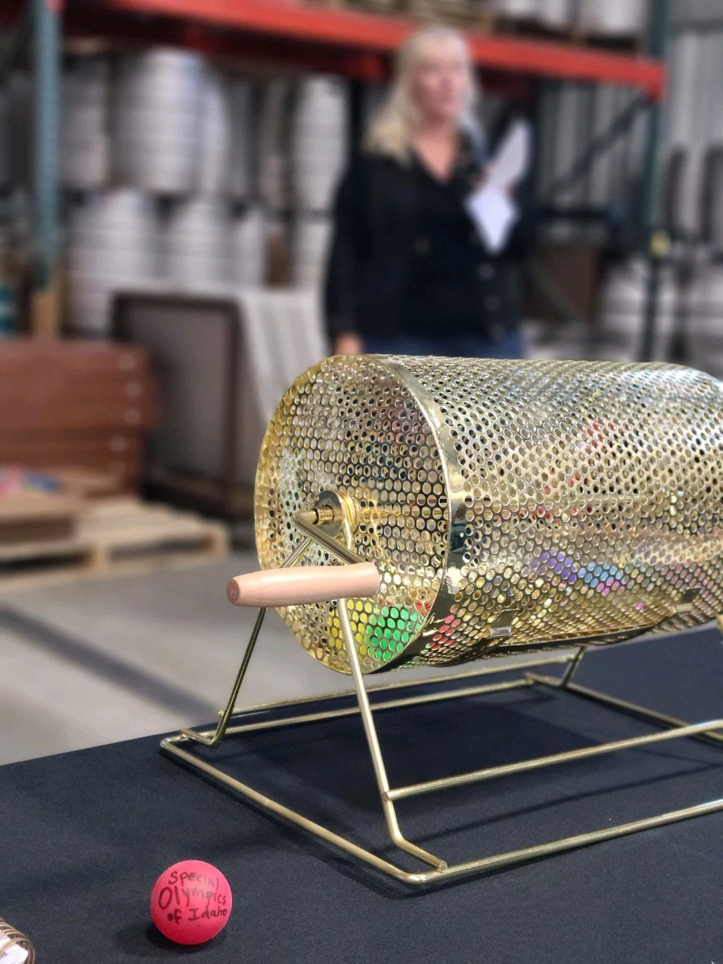 A golden raffle drum with a pink handle, placed on a dark table. In the background, a woman with blonde hair, wearing a black jacket, stands in a warehouse-like setting with shelves and rolls of material. A pink sticker on the table reads "Special Ol