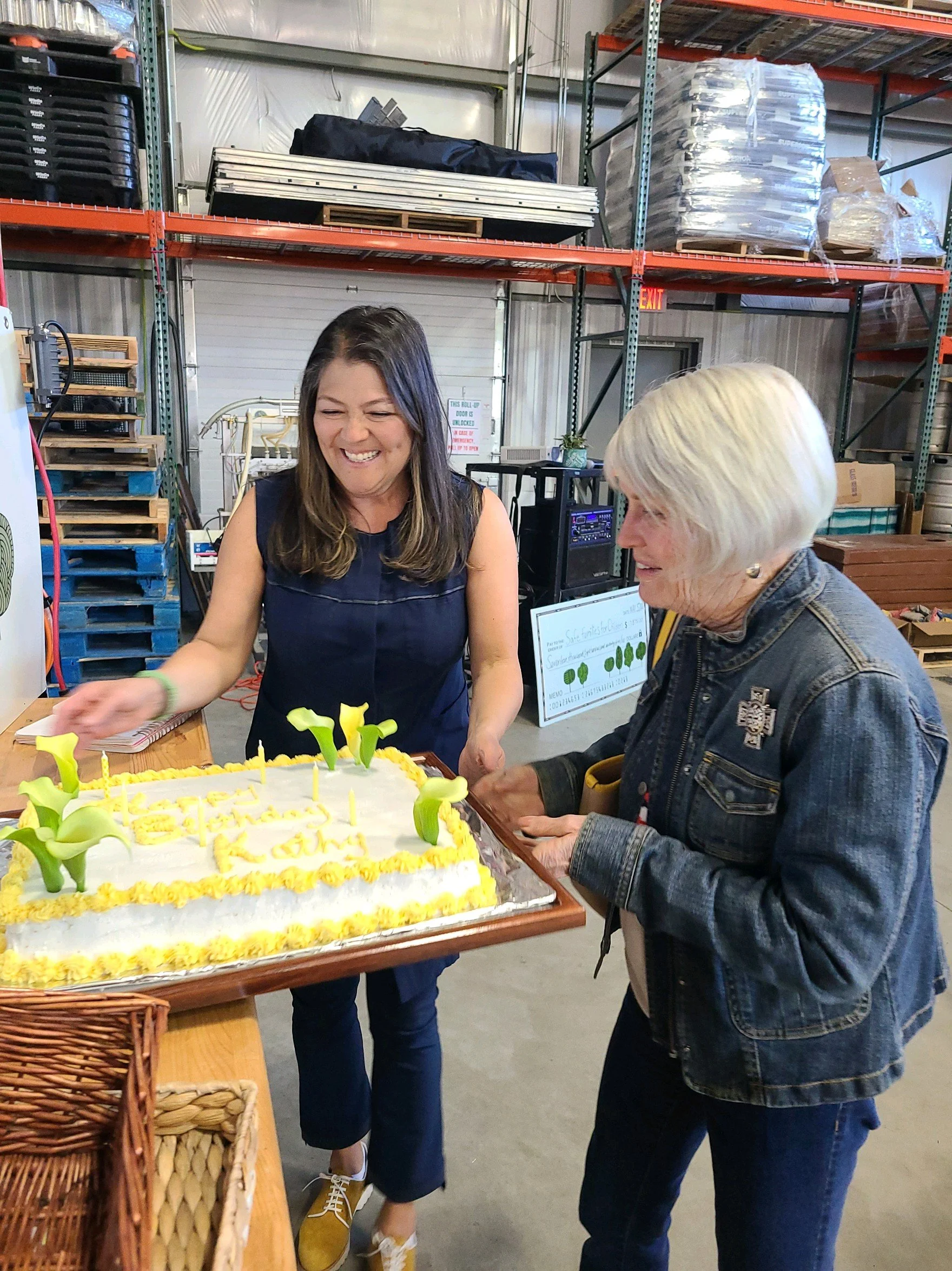 Two women are celebrating with a large cake, one woman is cutting the cake, and the other is holding the tray. They are in a warehouse or storage area with metal shelves and boxes in the background.