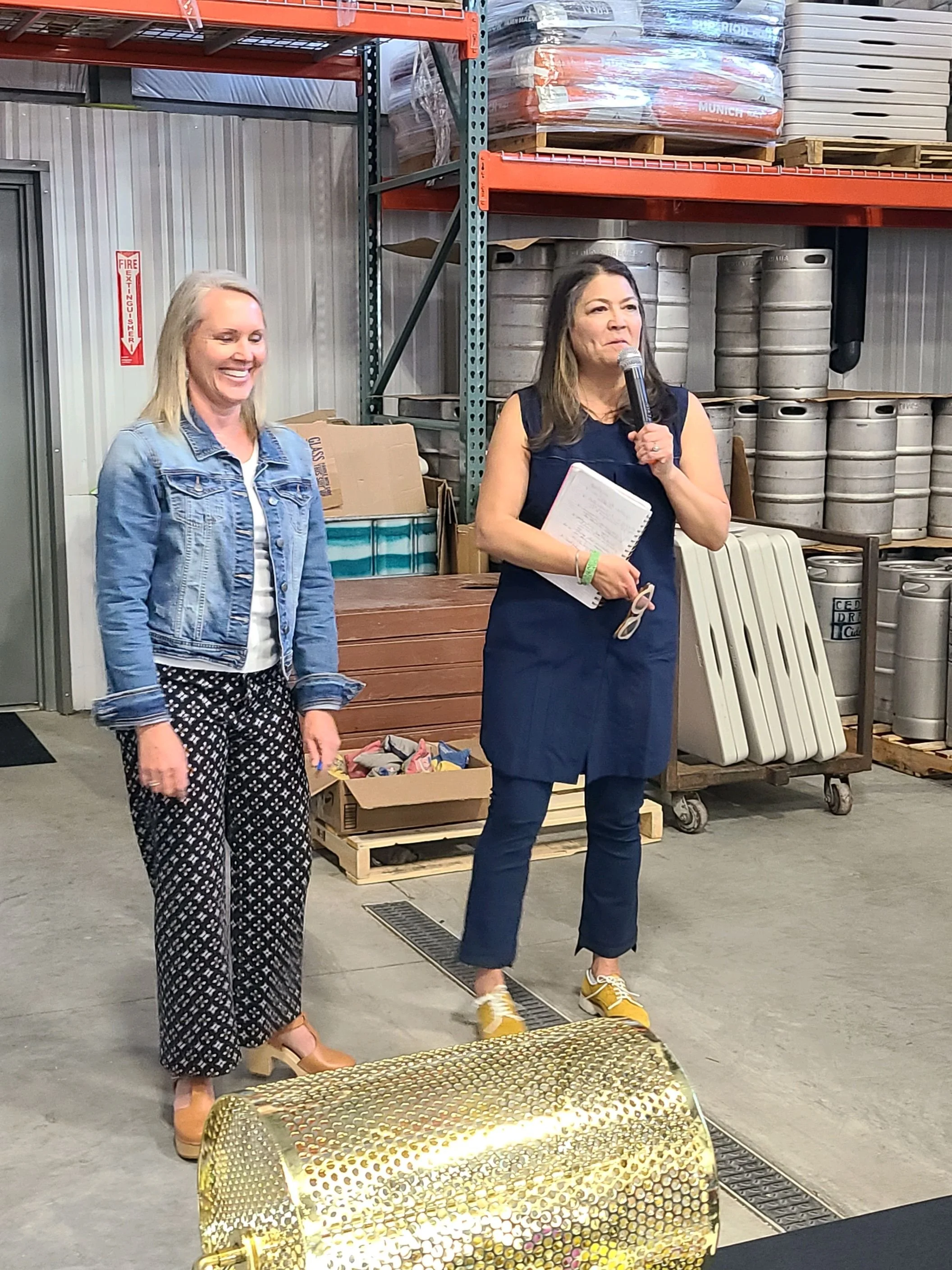 Two women standing in a warehouse, one holding a microphone and a notepad, the other smiling with hands by her sides. The warehouse is stocked with metal barrels, pallets, and boxes.