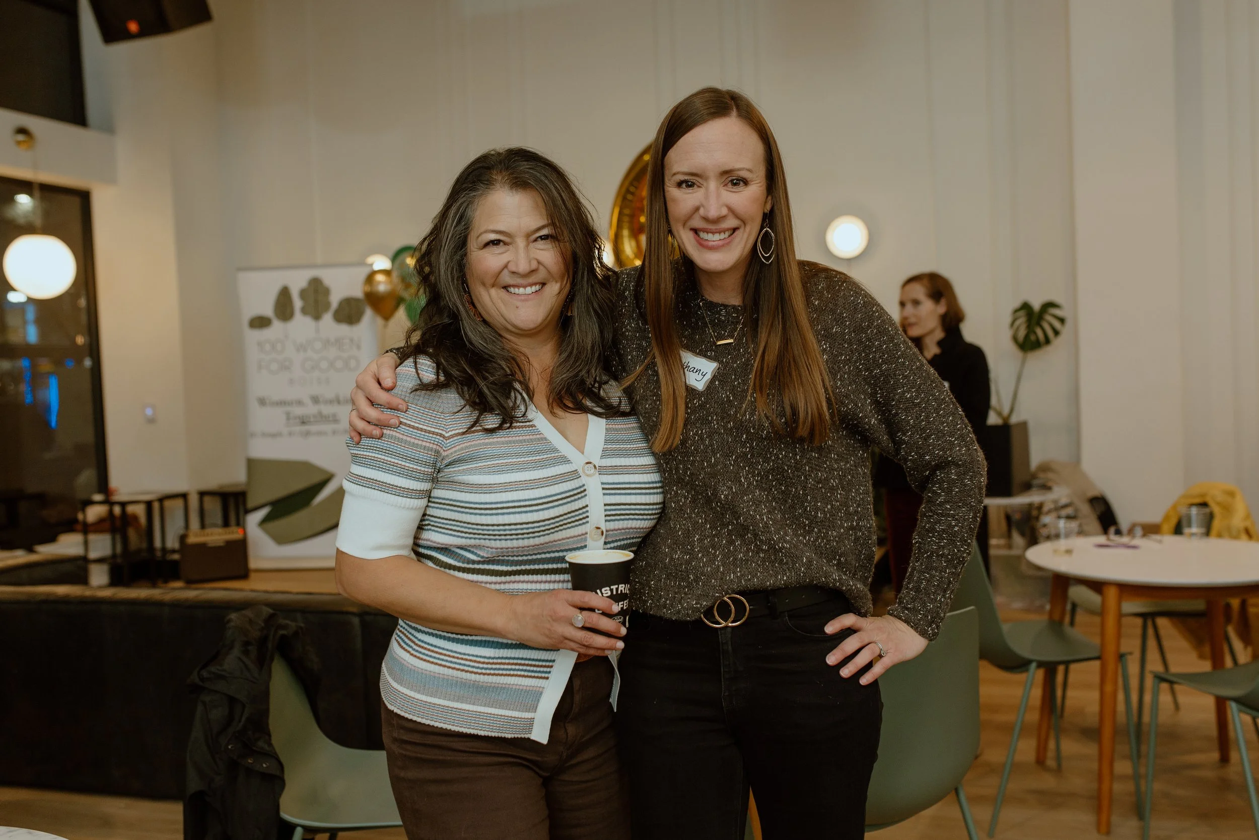 Two women smiling and posing together at an indoor event, with one holding a coffee cup, in a decorated room with tables and chairs.