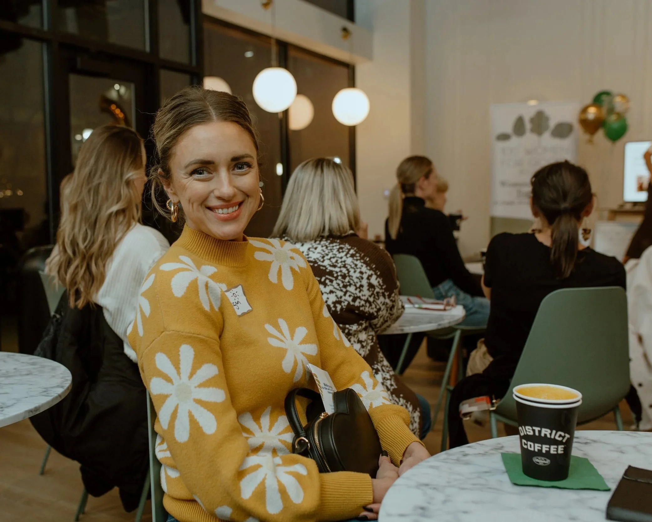 A woman with brown hair smiling, wearing a yellow sweater with white flower patterns, sitting at a marble table in a coffee shop or event space. There is a coffee cup with a green napkin on the table. Several other people are sitting at tables in the background.