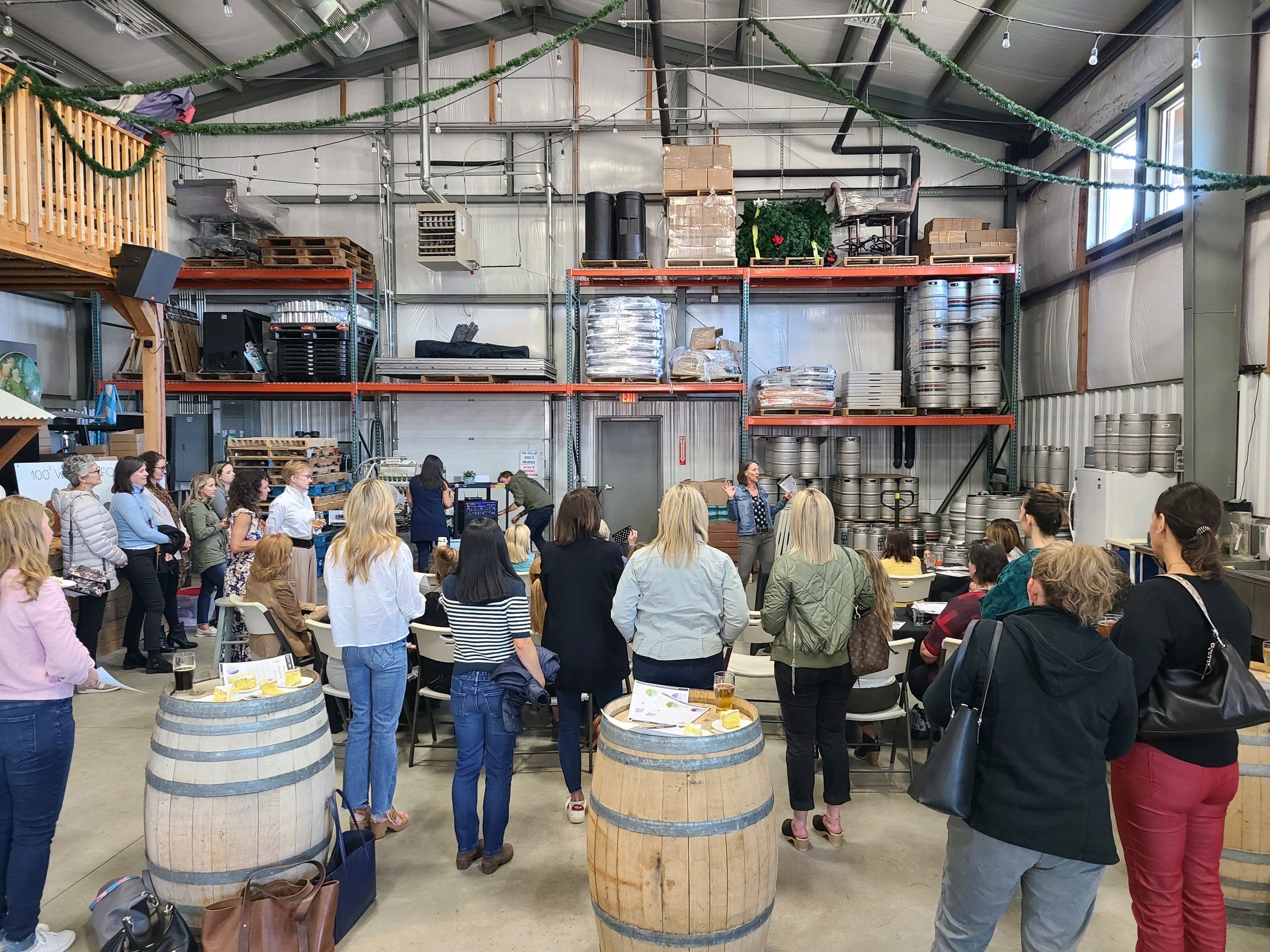 A group of people attending a presentation or tasting event inside a brewery or distillery, with stacks of kegs and shelves of supplies in the background.