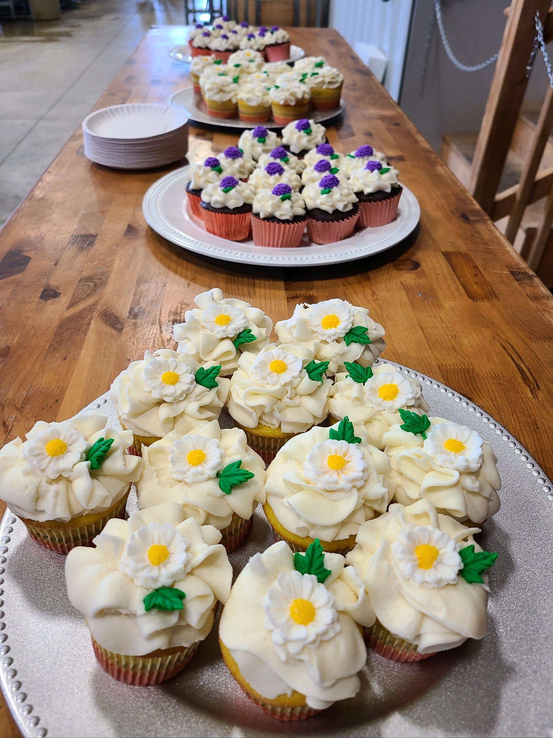 A variety of cupcakes decorated with white, purple, and yellow frosting, including daisy-shaped flowers, arranged on silver and white serving trays on a wooden table.