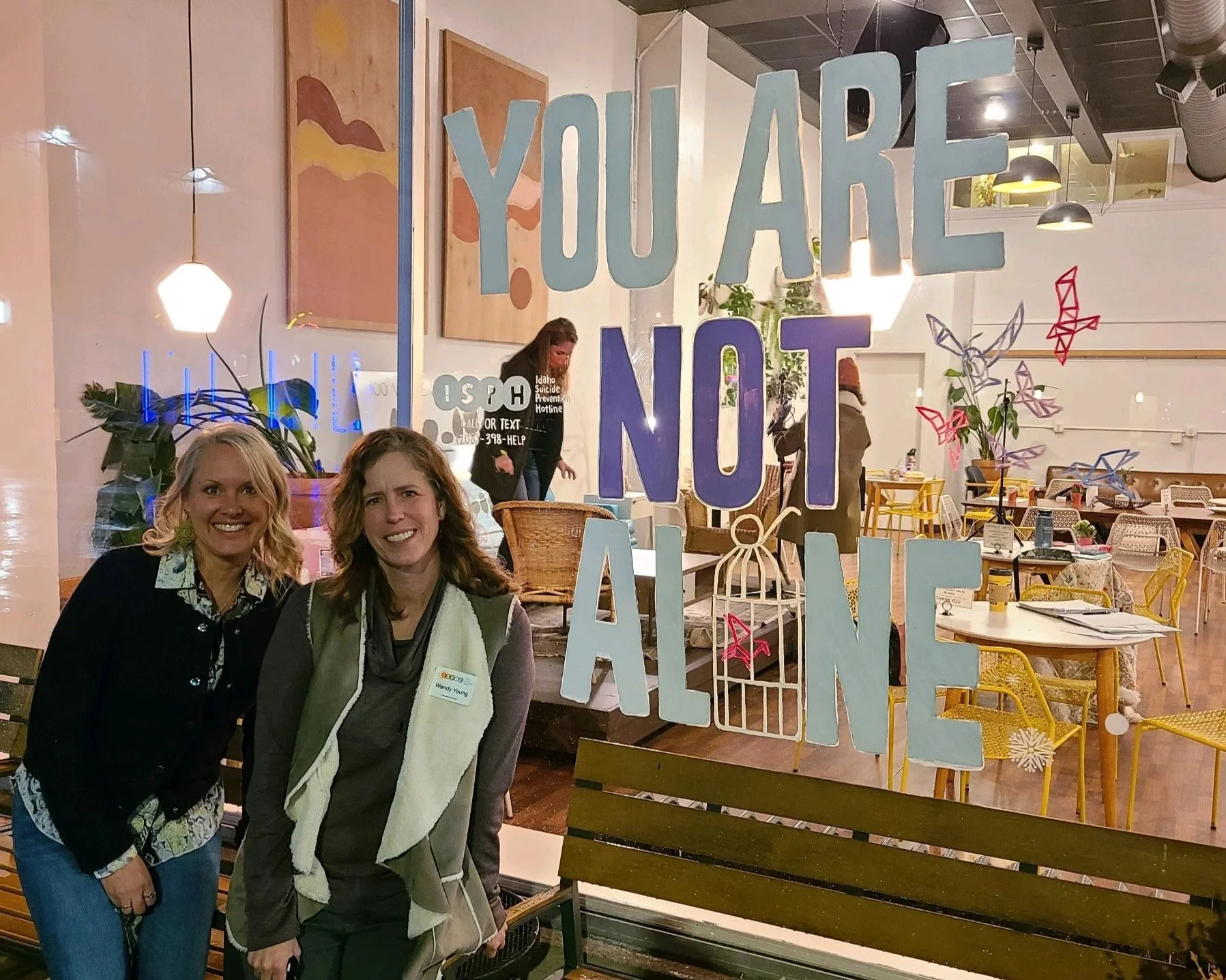 Two women standing in front of a glass window with colorful signs and art inside a cafe or restaurant. The sign on the window reads, 'YOU ARE NOT ALONE' in large colorful letters.