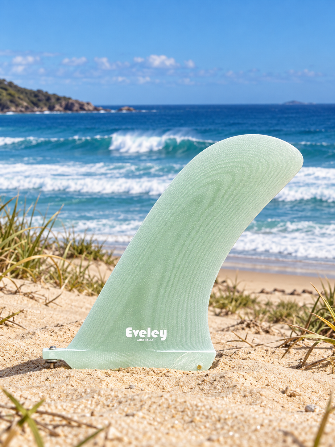Natural translucent Eveley Santa Monica fin standing in dune sand with breaking surf behind it.