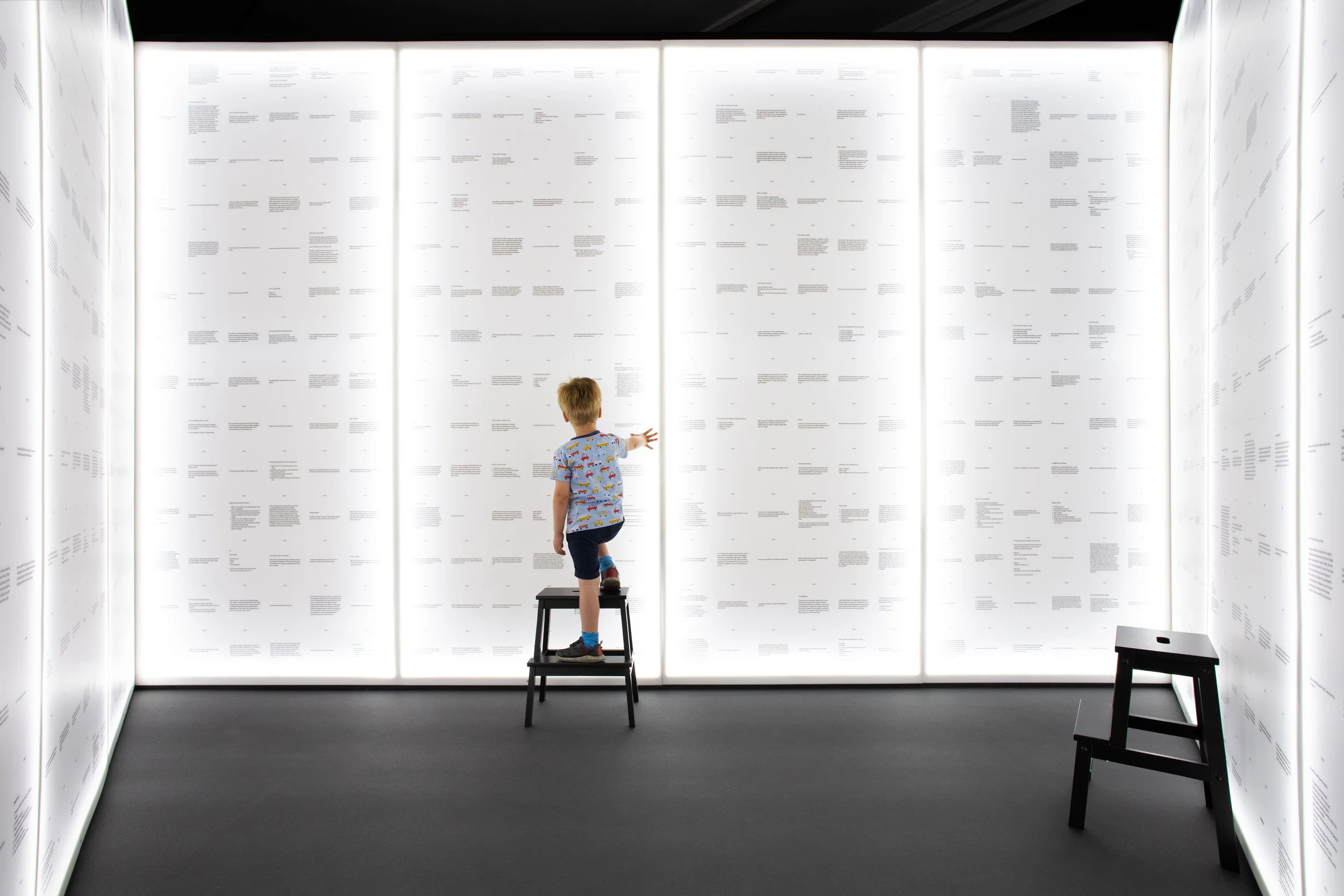A young boy in a colorful shirt and shorts standing on a small stool, pointing at a large illuminated wall of text in an art or museum exhibit.