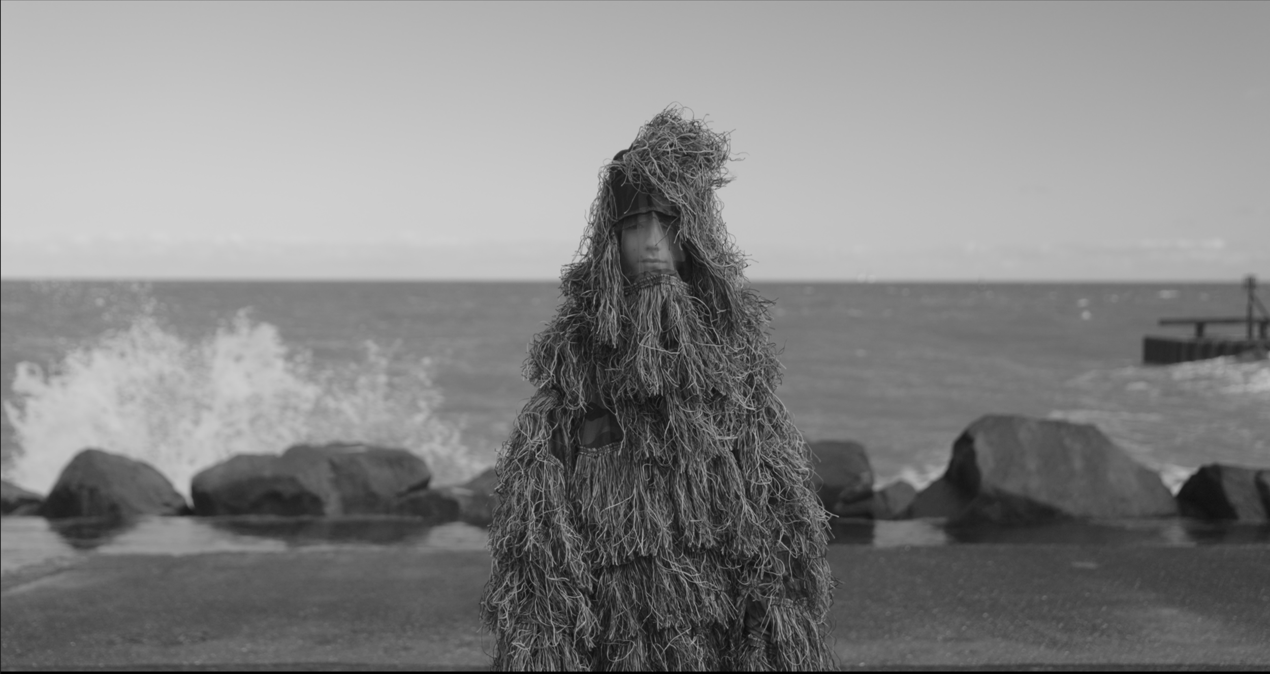 Person dressed in a shaggy, ghillie-style costume standing on a beach with rocks and ocean in the background, in black and white.