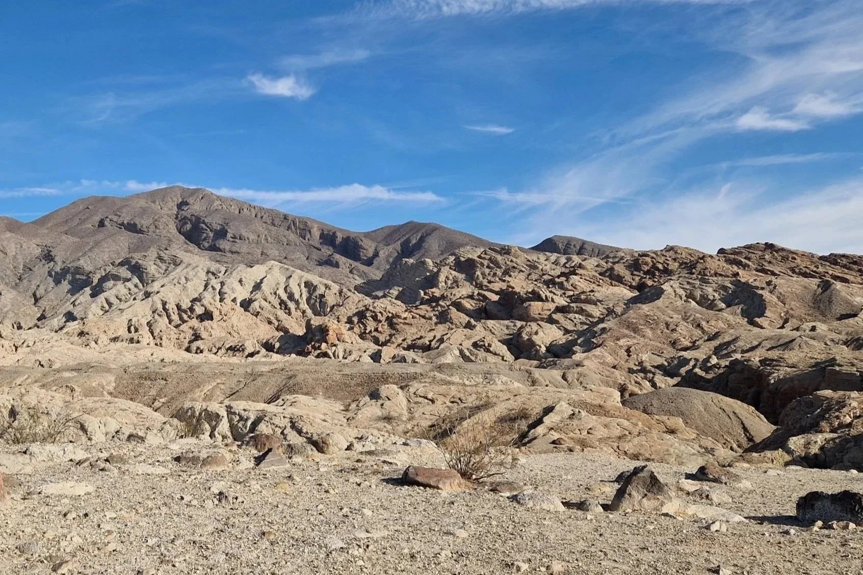 Desert landscape with rocky terrain, mountains in the background, and a partly cloudy blue sky.
