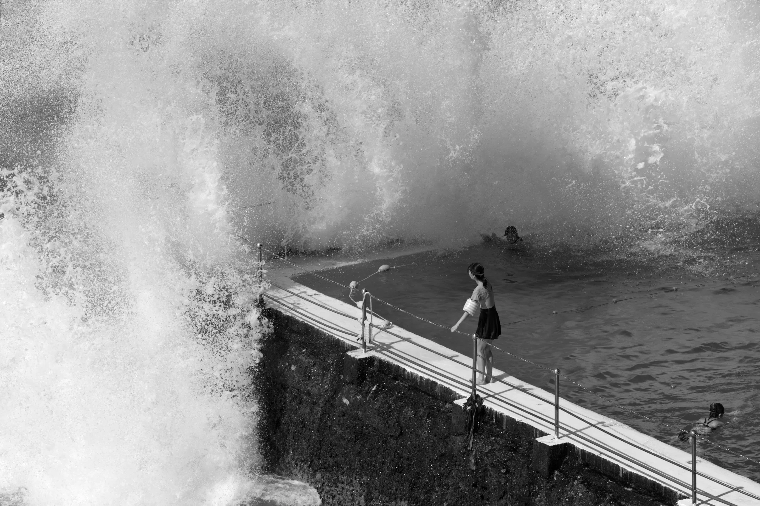 A young girl standing on a narrow fence at a seaside walkway while large waves crash against the rocks, with some people swimming in the water in the background.