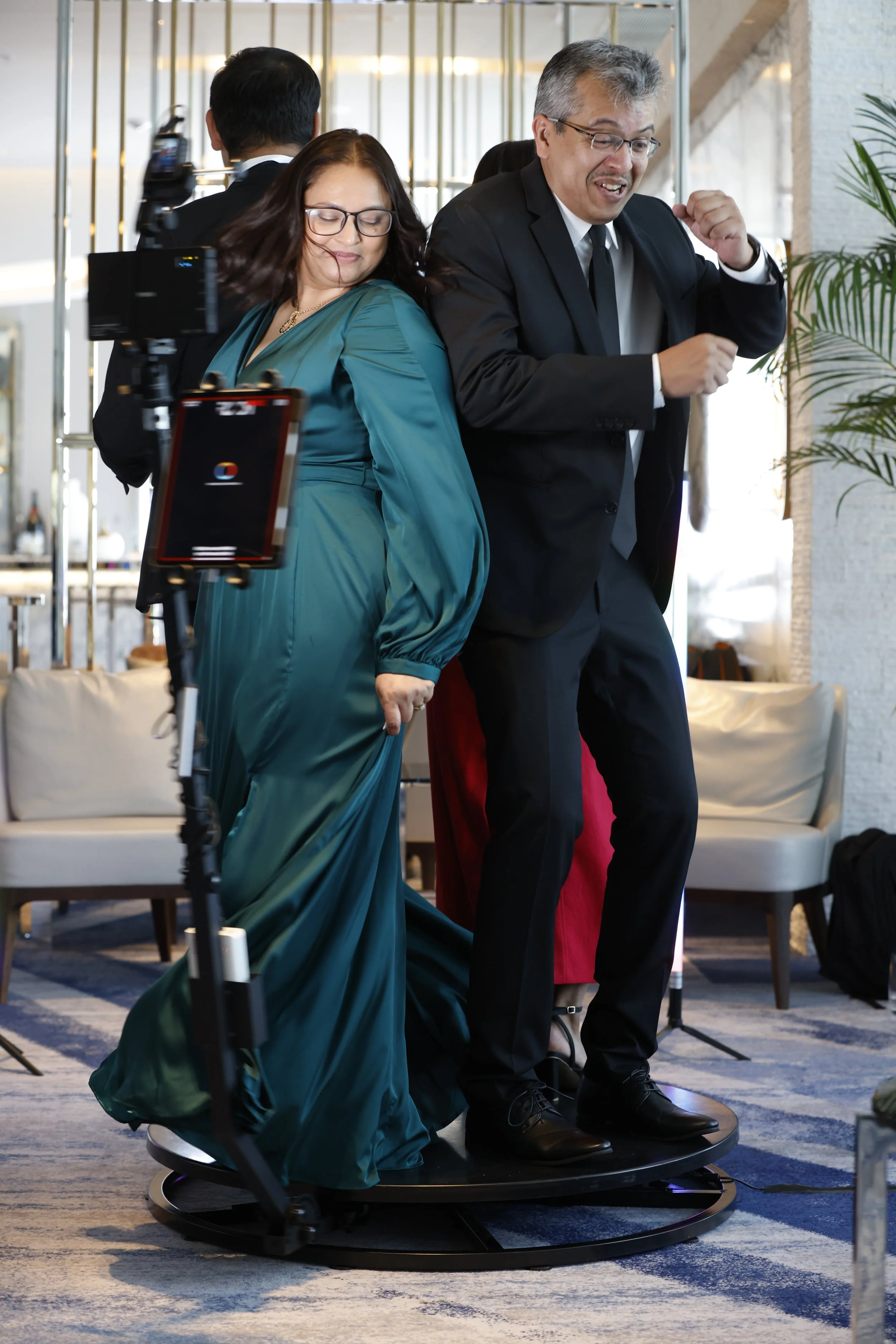 Two people on a revolving platform dancing at a social event, with one woman in a teal gown and glasses, and one man in a black suit with glasses, both smiling and enjoying the moment.
