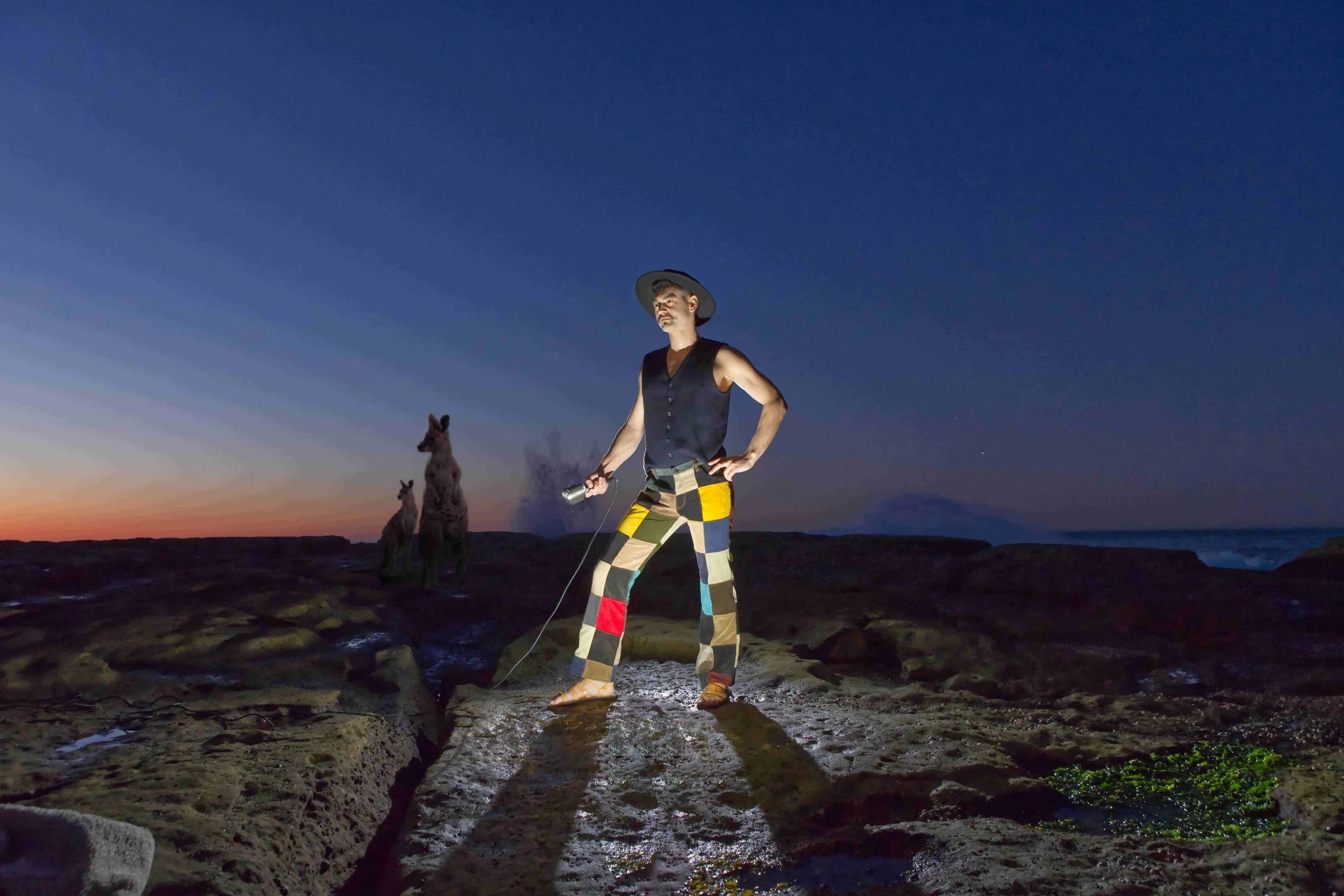 A man stands on rocks at sunset, holding a flashlight, wearing a sleeveless vest and colorful patchwork pants, with two giraffes in the background and the ocean visible to the right.