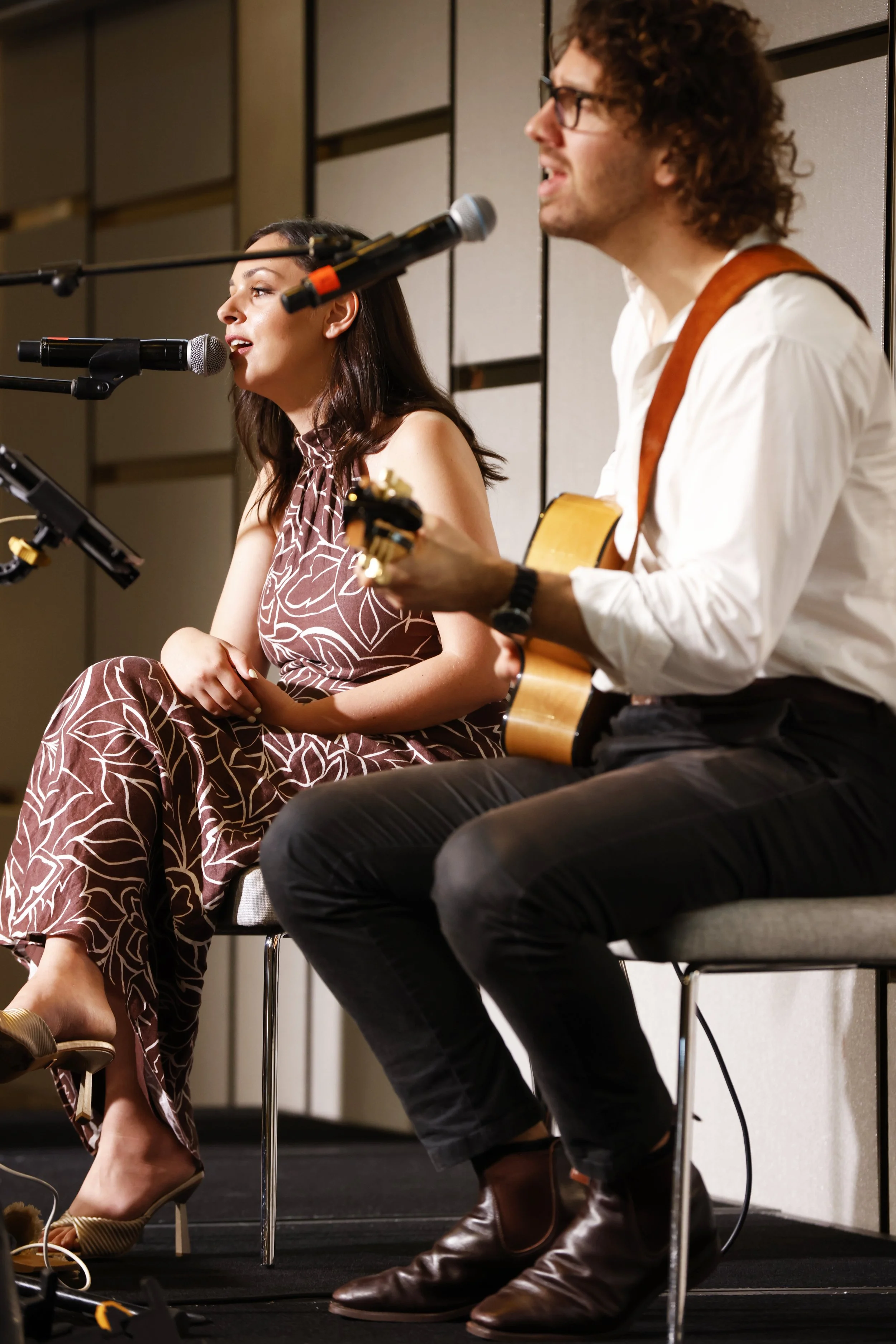 A woman and a man performing music on stage, with the woman singing into a microphone and the man playing an acoustic guitar.