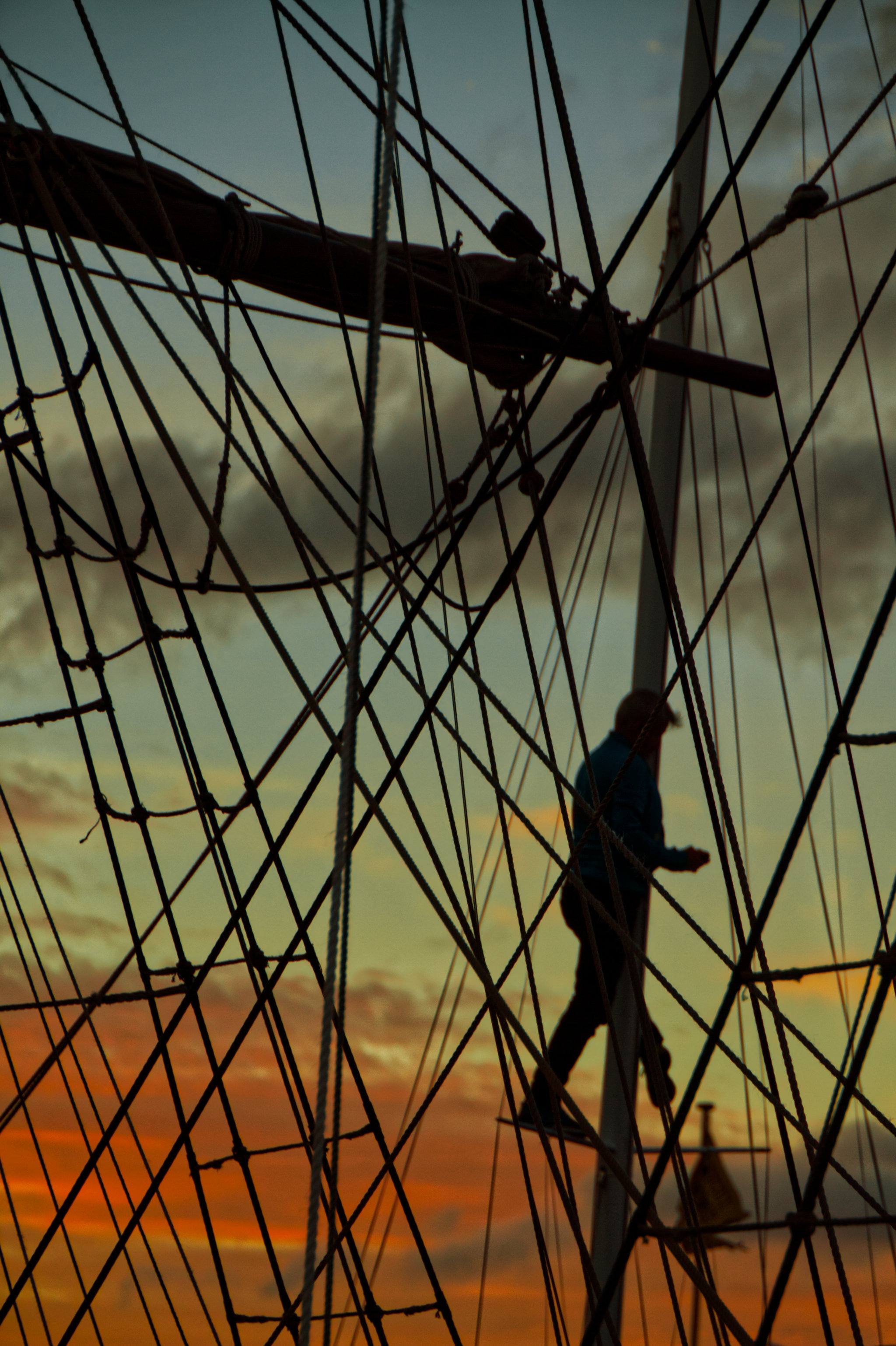 Silhouette of a person climbing rigging on a tall ship at sunset, with ropes and mast visible.