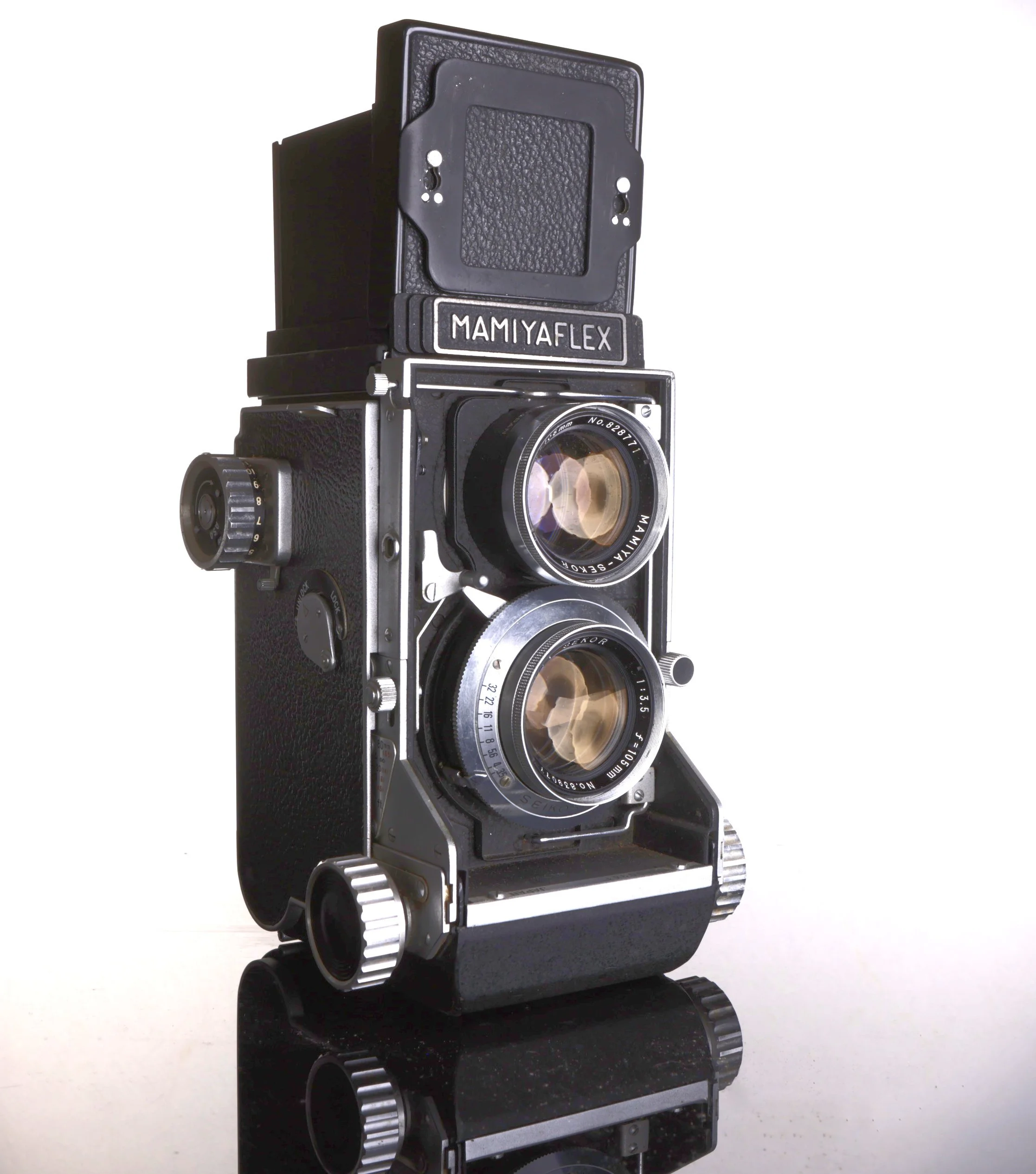 Vintage Mamiya Flex twin-lens reflex camera with black body and silver accents reflected on a black glossy surface.
