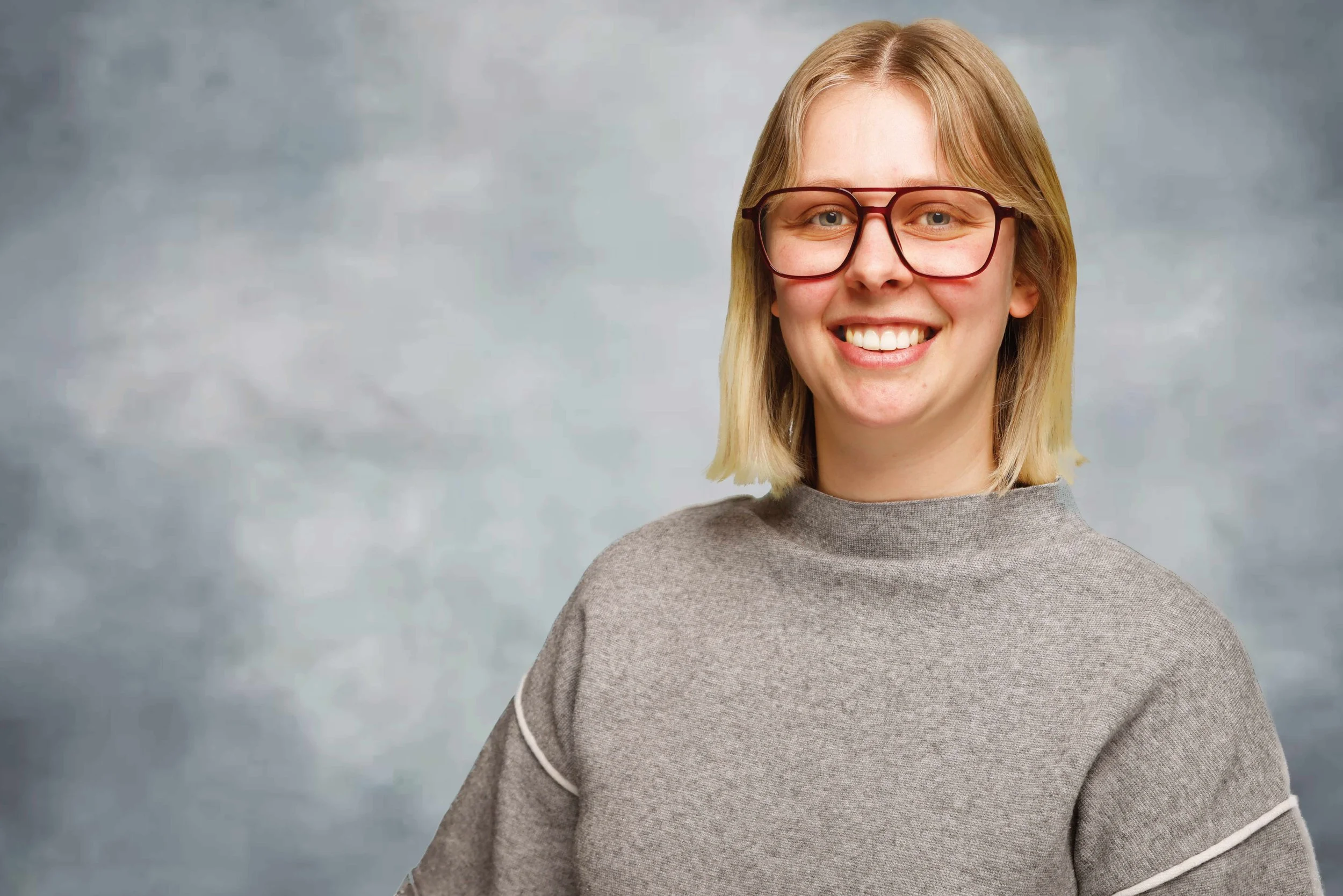 A young woman with shoulder-length blond hair and glasses smiling, wearing a gray sweater, standing against a blurry gray background.