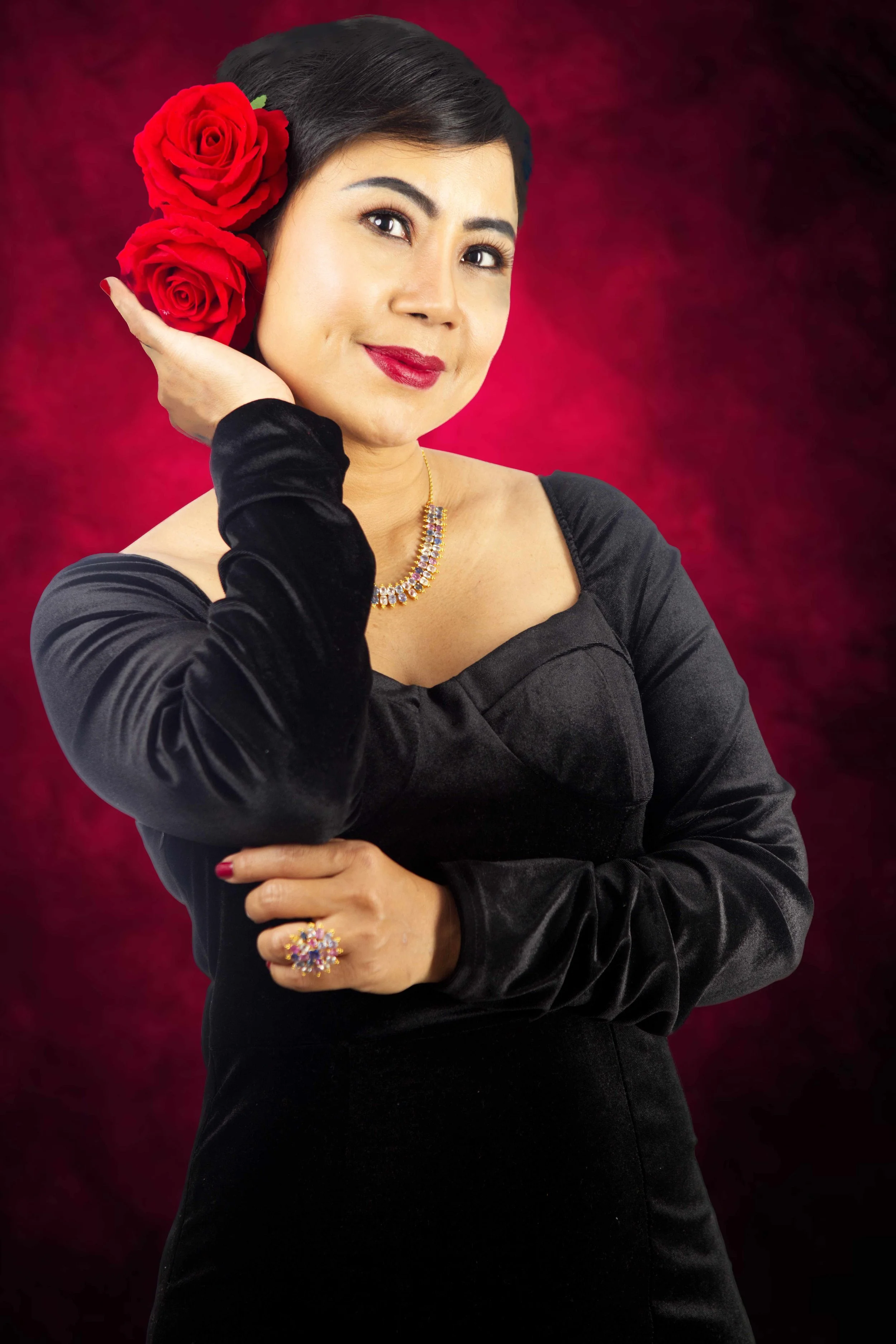 Woman with dark hair and red lipstick, wearing a black dress, gold jewelry, holding a cluster of three red roses to her head, against a red and black background.