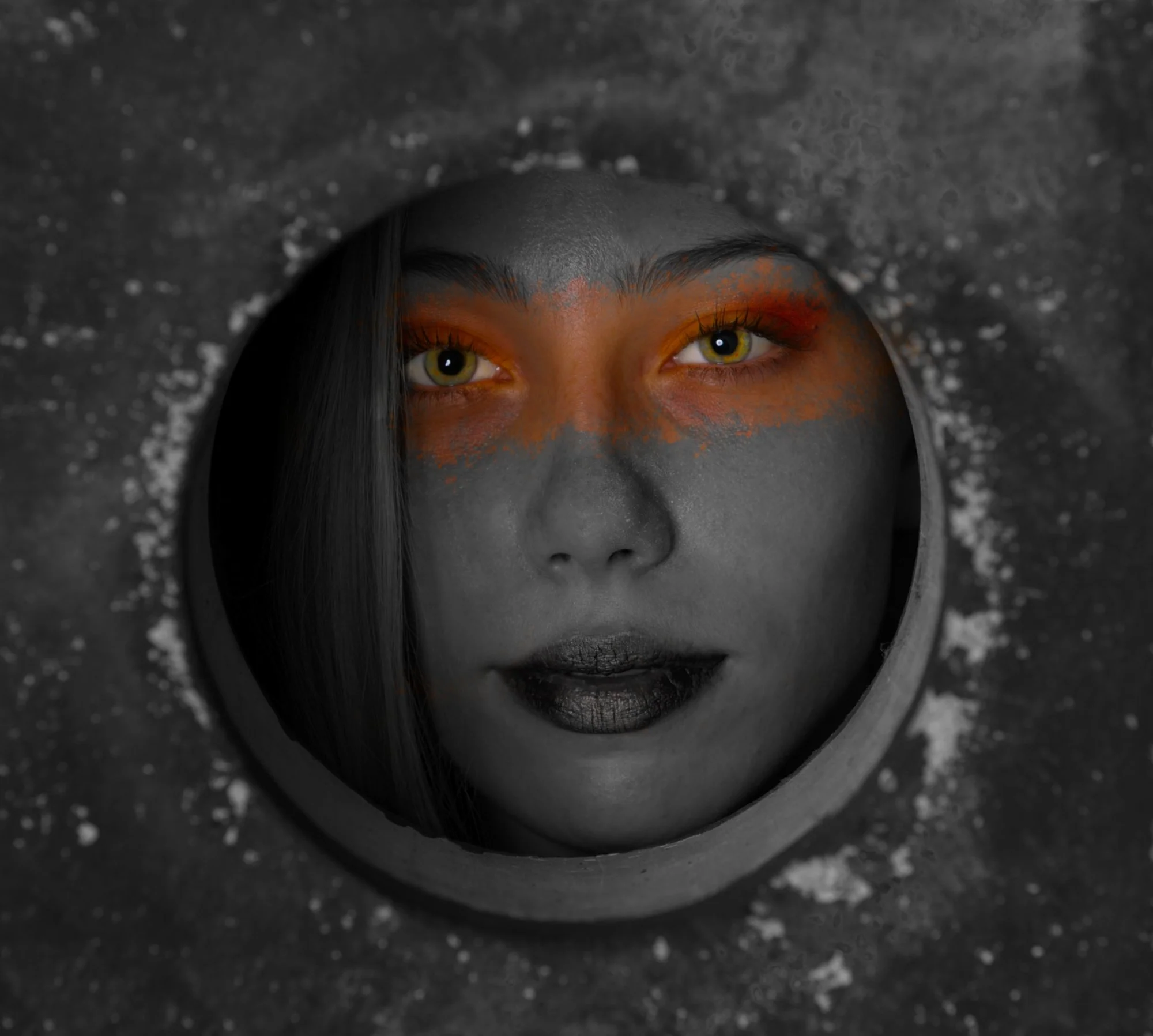 A person's face is seen through a circular hole in a textured dark surface. The person's face is in black and white, with bright orange eyeshadow and yellow-green eyes. Their lips are painted black, and they have long dark hair.